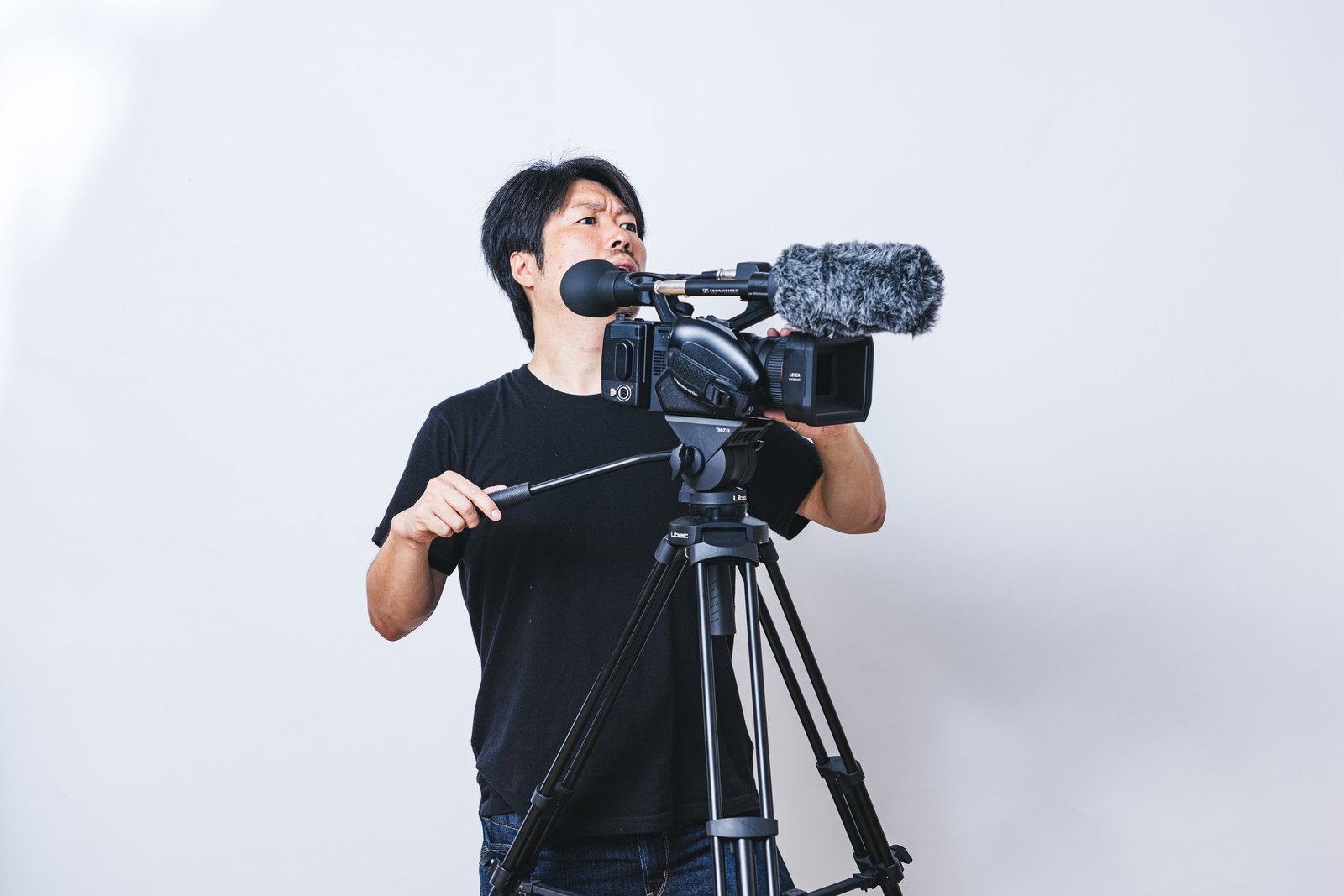 黒いTシャツを着たマスク姿の男性カメラマンが業務用ビデオカメラを操作している［モデル：瀧野恵太］