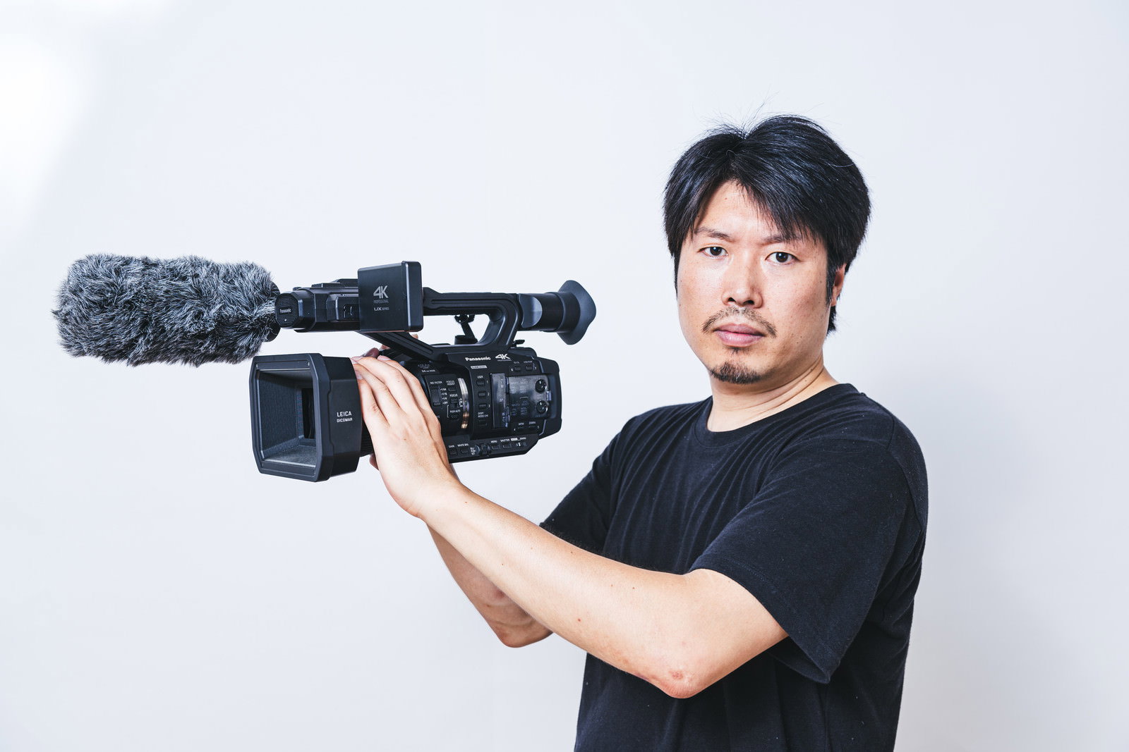 業務用ハンディビデオカメラを肩に構えて撮影する黒いTシャツの男性［モデル：瀧野恵太］