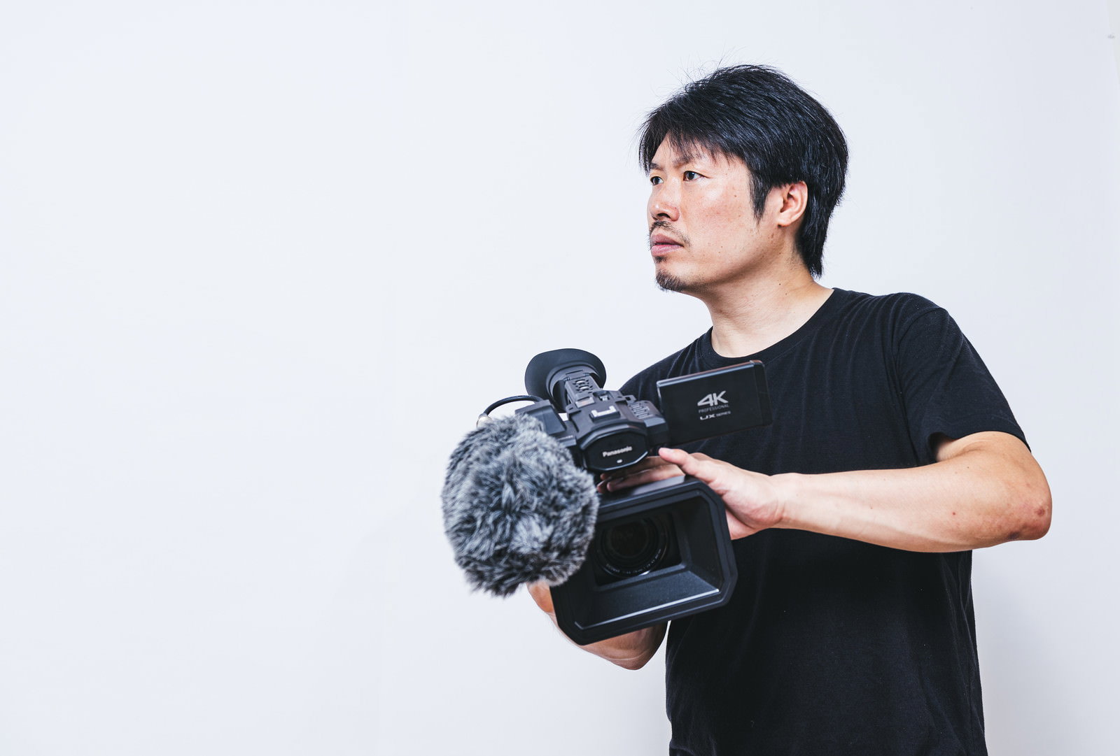 ハンディカメラを両手で構えて撮影準備をする黒いTシャツの男性カメラマン［モデル：瀧野恵太］