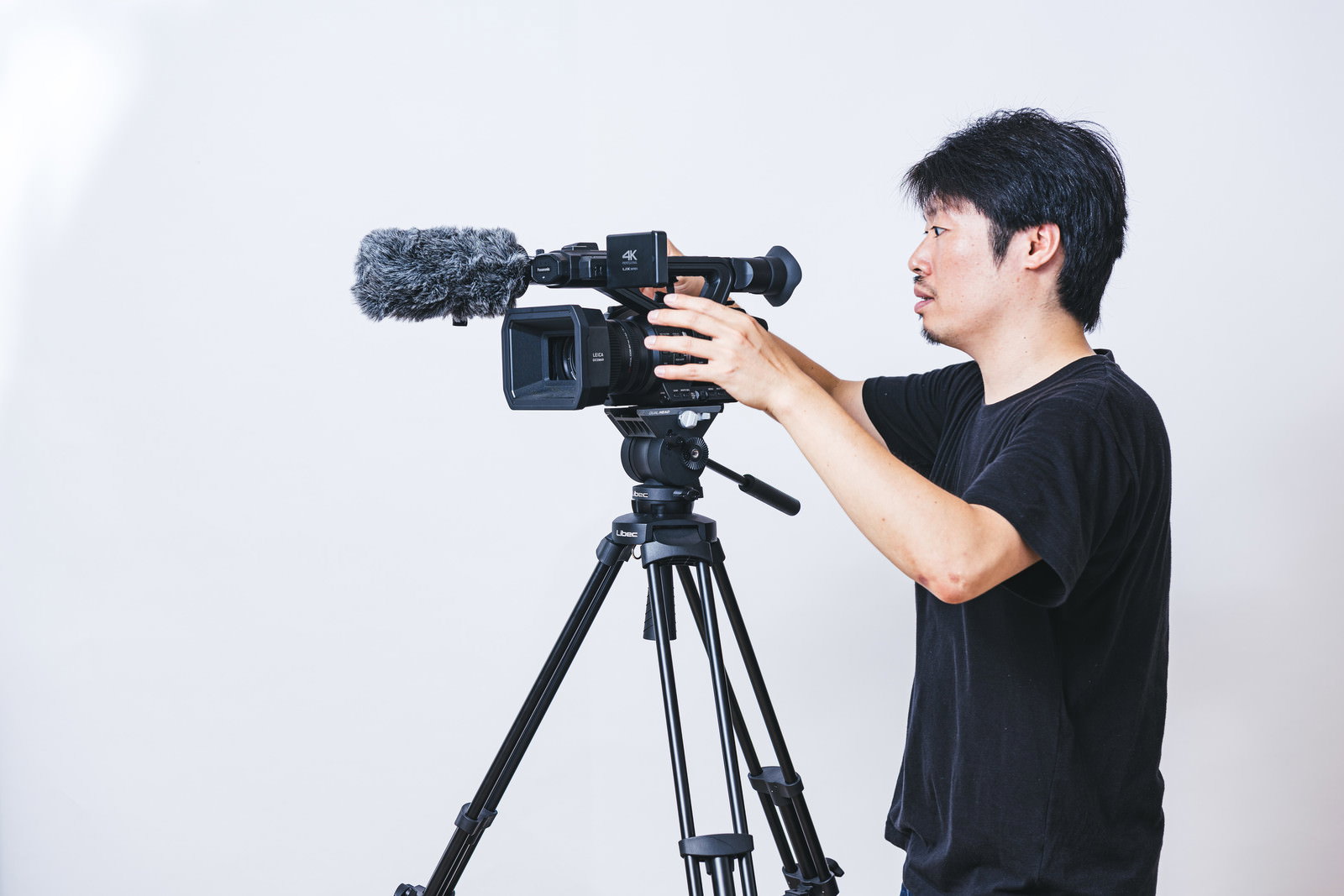 三脚に設置された業務用4Kビデオカメラを操作する黒いTシャツの男性カメラマン［モデル：瀧野恵太］