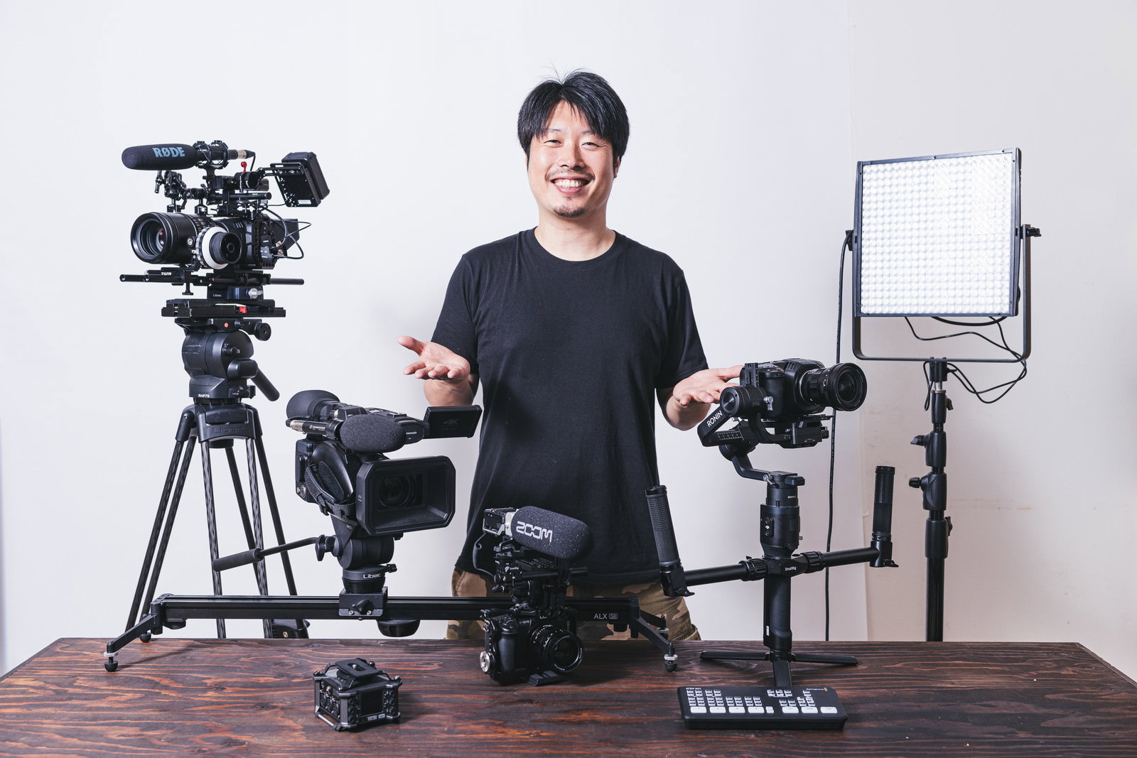 笑顔で手を広げながら動画撮影機材を紹介する黒いTシャツの男性［モデル：瀧野恵太］