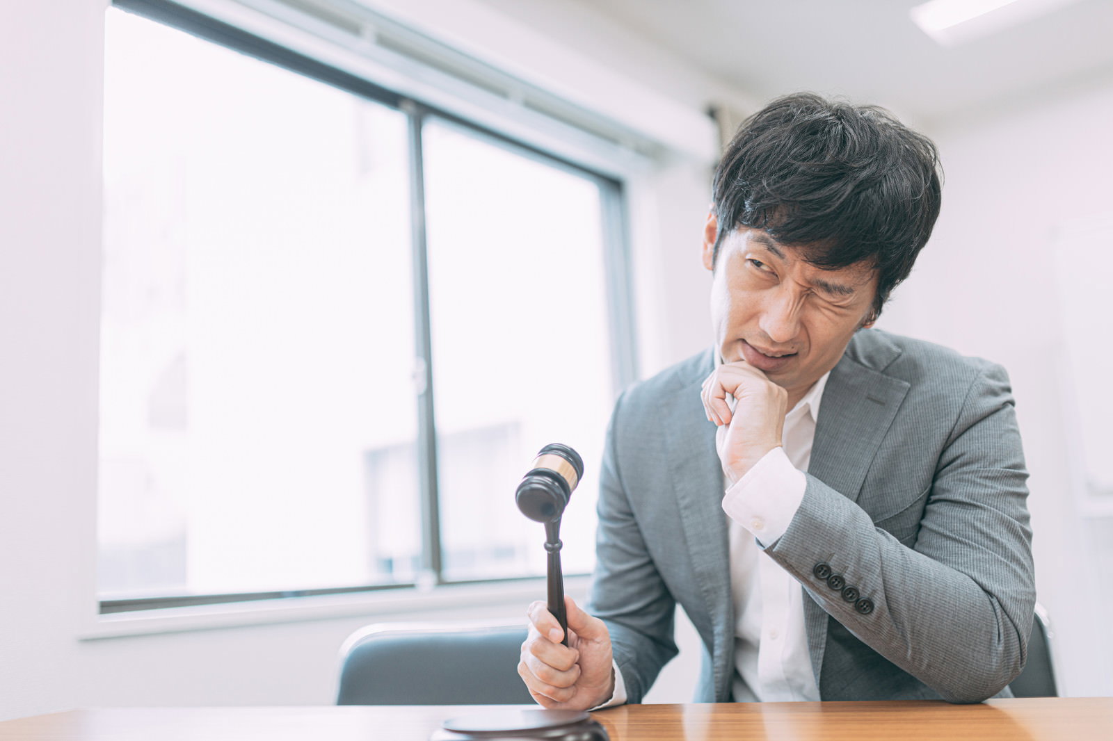 ハンマーを持って頬杖をつき考え込むグレーのスーツの男性［モデル：大川竜弥］
