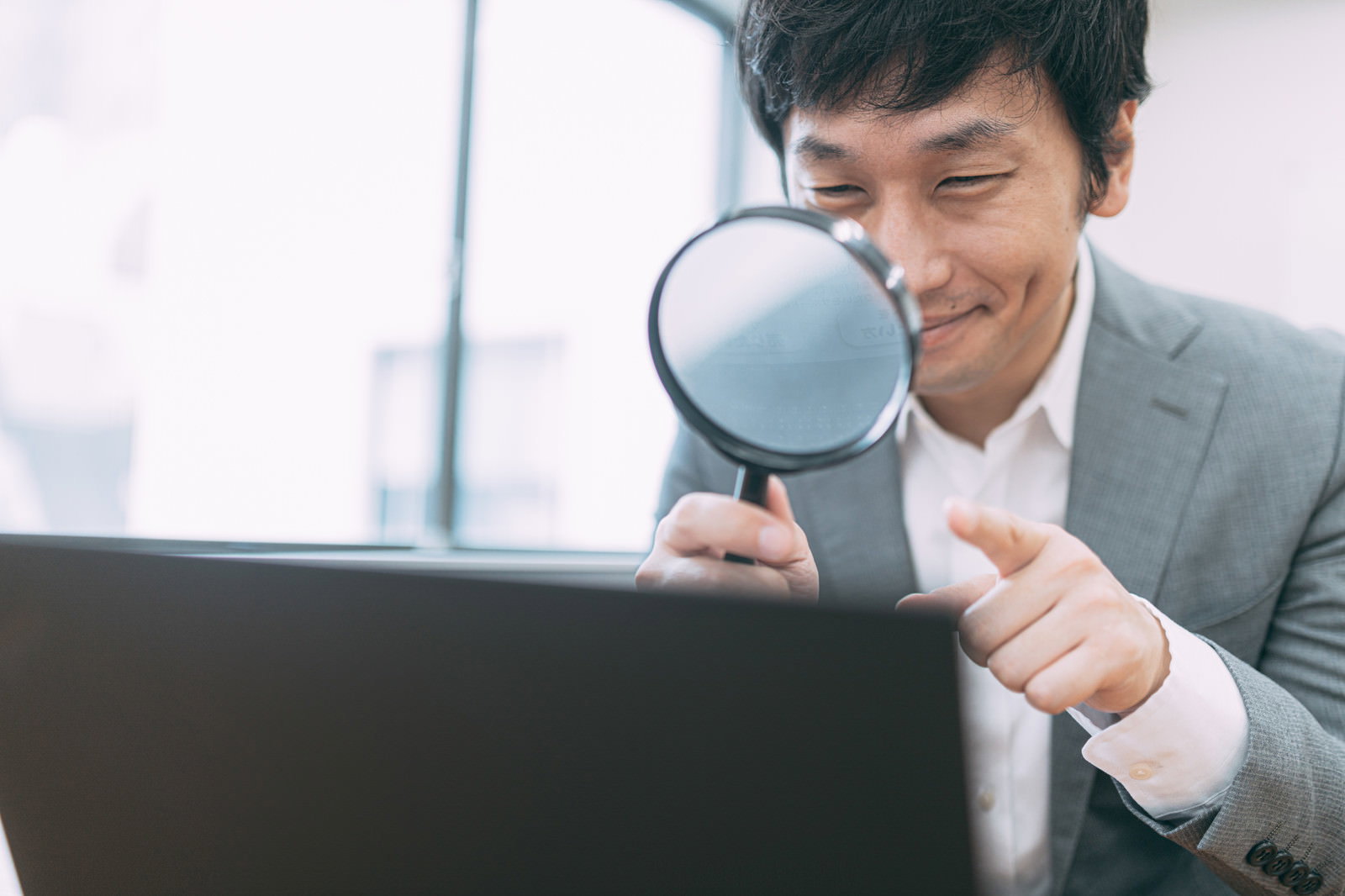 虫眼鏡を持ちながらパソコンで調査作業をするスーツ姿の男性［モデル：大川竜弥］