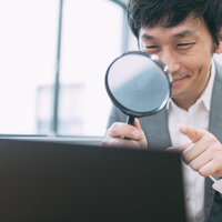 虫眼鏡でパソコンの調査に没頭するスーツ姿の男性の写真