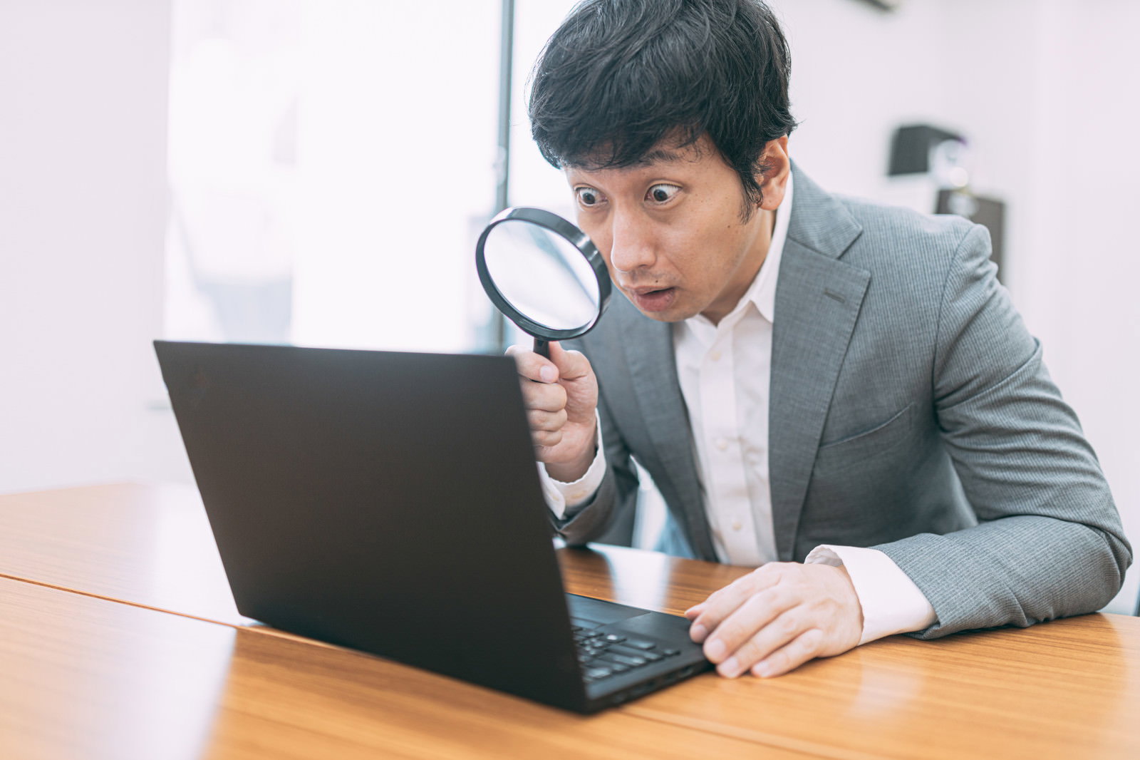 虫眼鏡を使ってノートパソコンの画面を詳しく調査している男性［モデル：大川竜弥］