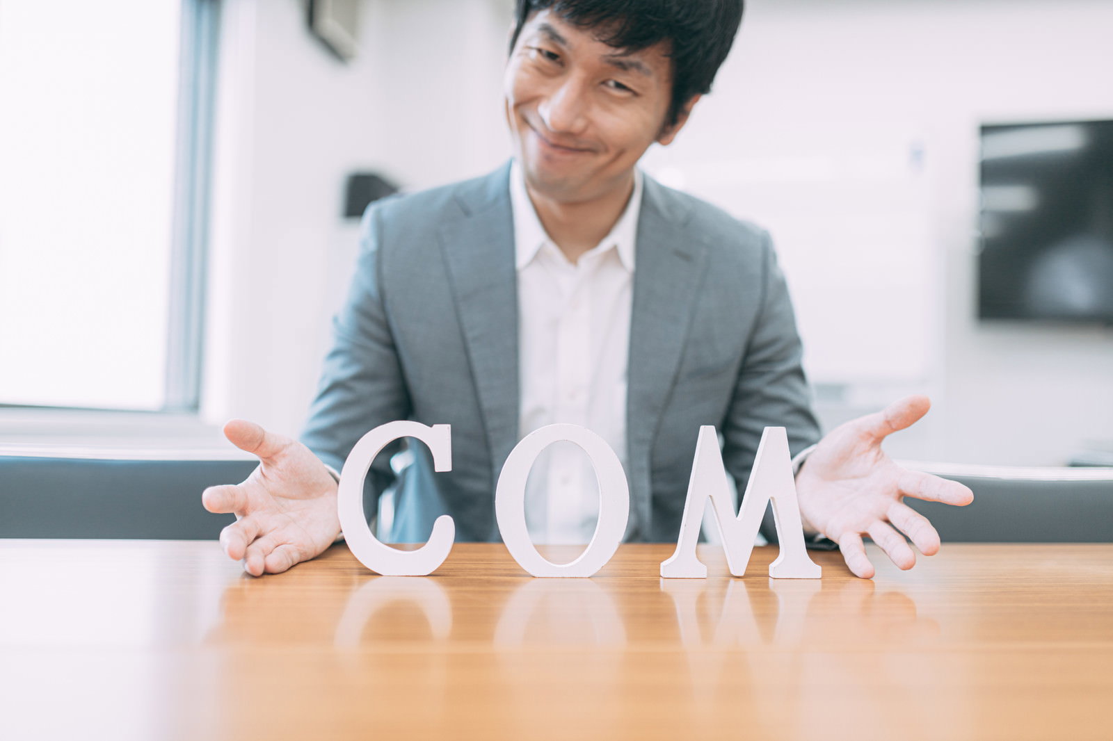 COMの文字を両手で示すグレーのスーツを着たビジネスマン［モデル：大川竜弥］