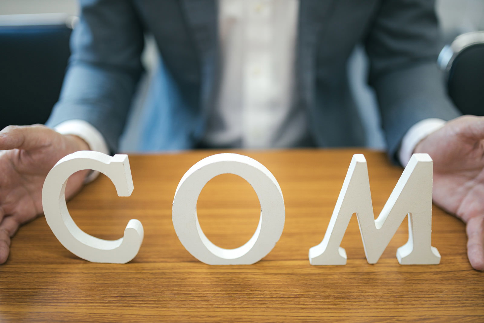 白い立体文字の「COM」を両手で支えながら説明する、スーツ姿の男性。木製テーブルの上での説明風景［モデル：大川竜弥］
