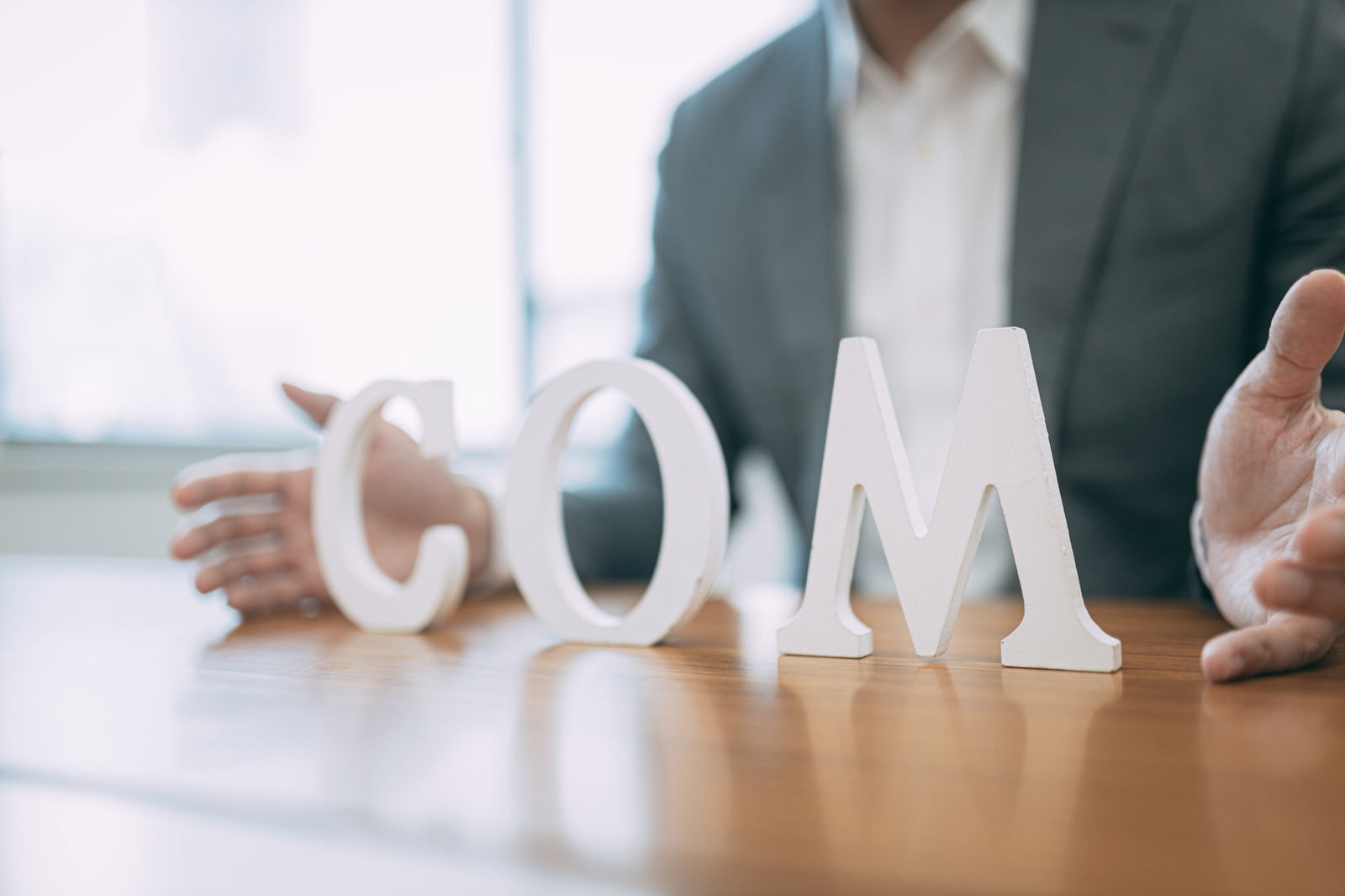 木製デスクの上でCOMの立体文字を指差すスーツ姿の男性［モデル：大川竜弥］