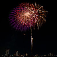 夜空に打ち上がる赤と紫の花火と夜景の競演の写真