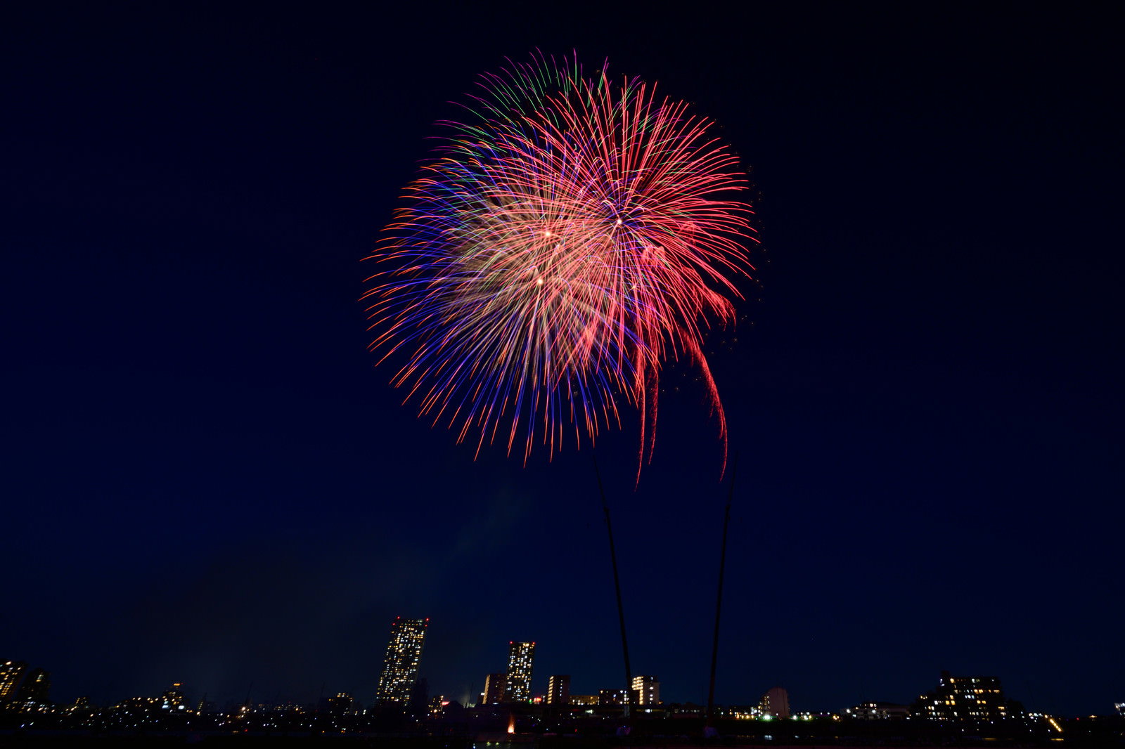 都会の夜空に色とりどりの花火が打ち上げられ、手前にビル群のシルエットが見える