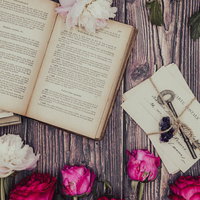 色褪せたピンクのバラと古書、手紙が並ぶマット上の静物の写真