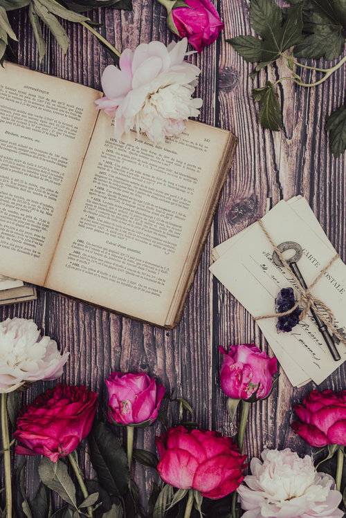 色褪せたピンクのバラと古書、手紙が並ぶマット上の静物