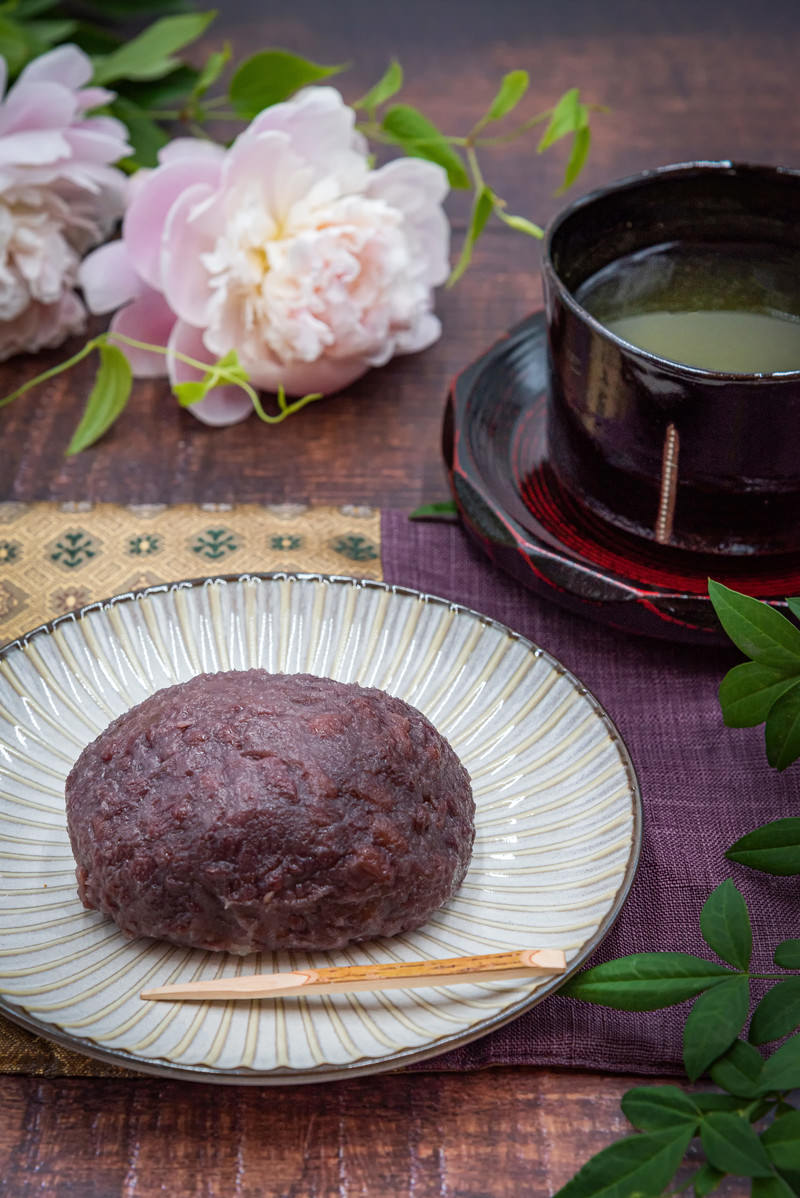 白い皿のおはぎと茶碗に入ったお茶、淡いピンクの牡丹の花が木のテーブルに配置された和の食卓