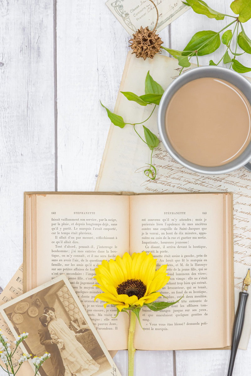 開かれた洋書の上に黄色い向日葵が置かれ、ティーカップと手紙が添えられた構図