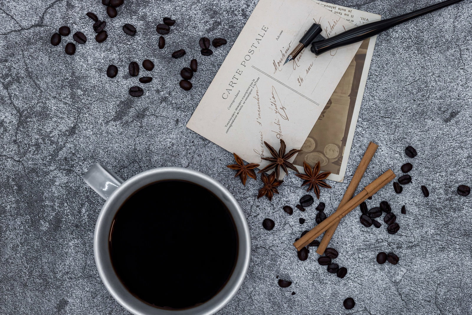 グレーの背景にコーヒーカップと散らばるコーヒー豆、絵葉書、万年筆が配置された写真