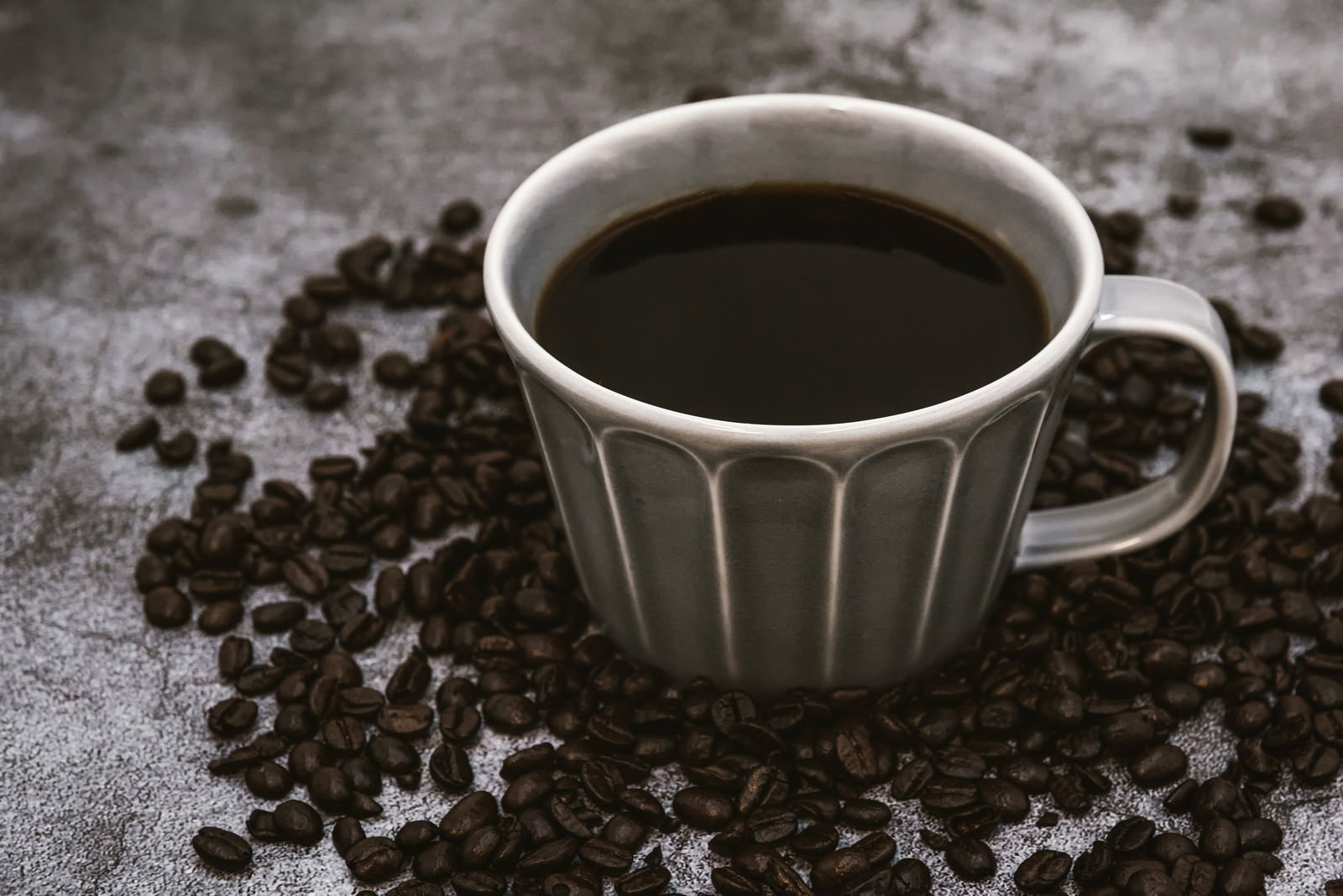 白いストライプ柄のマグカップに入ったブラックコーヒーと周囲に散らばった焙煎コーヒー豆