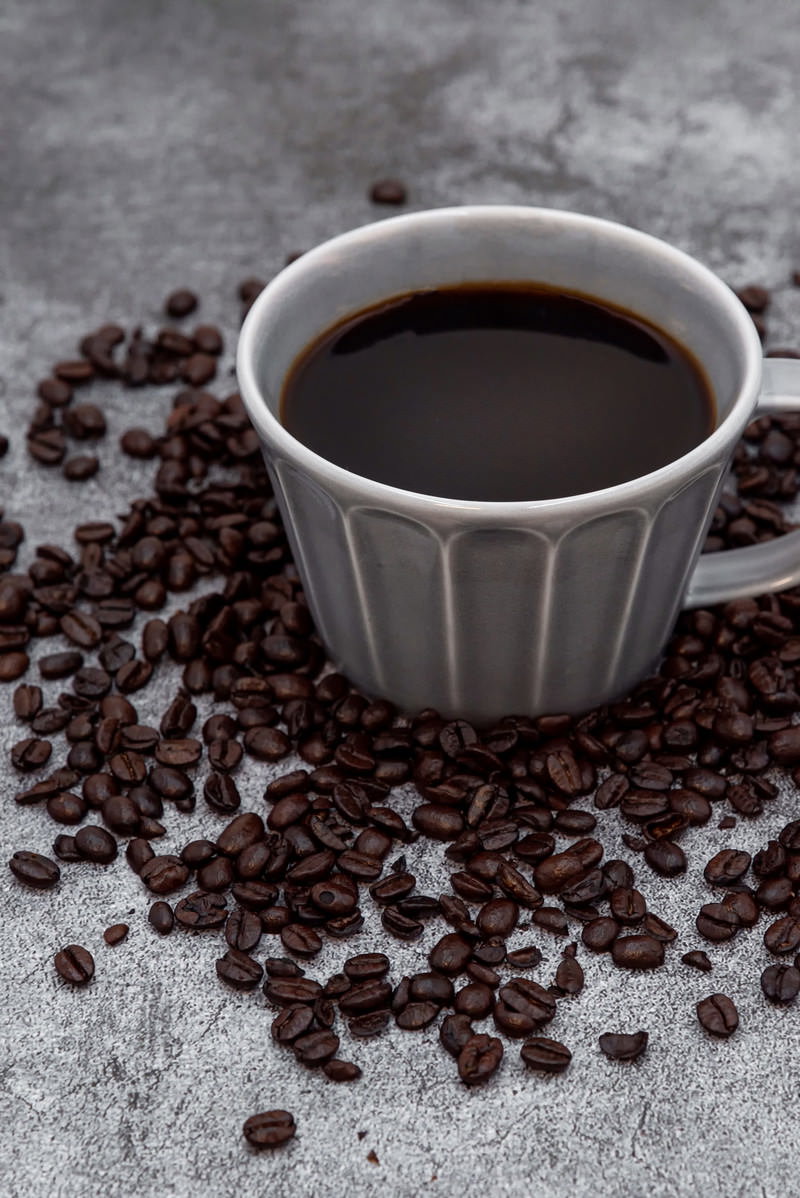 グレーのリブ付きカップに入ったブラックコーヒーとその周りに散らばったコーヒー豆