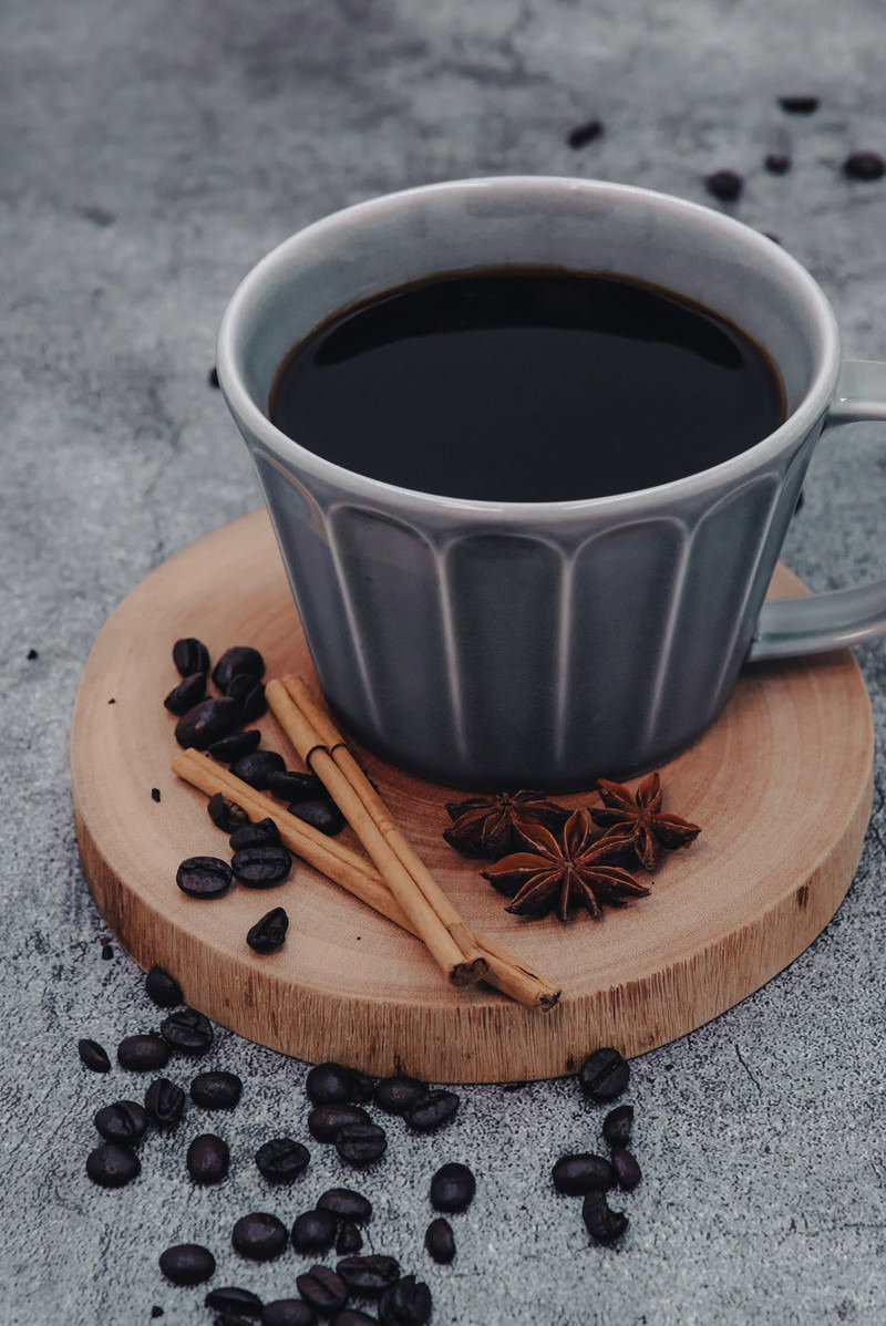 木製コースターの上に置かれたグレーのコーヒーカップとシナモンスティック、散らばったコーヒー豆