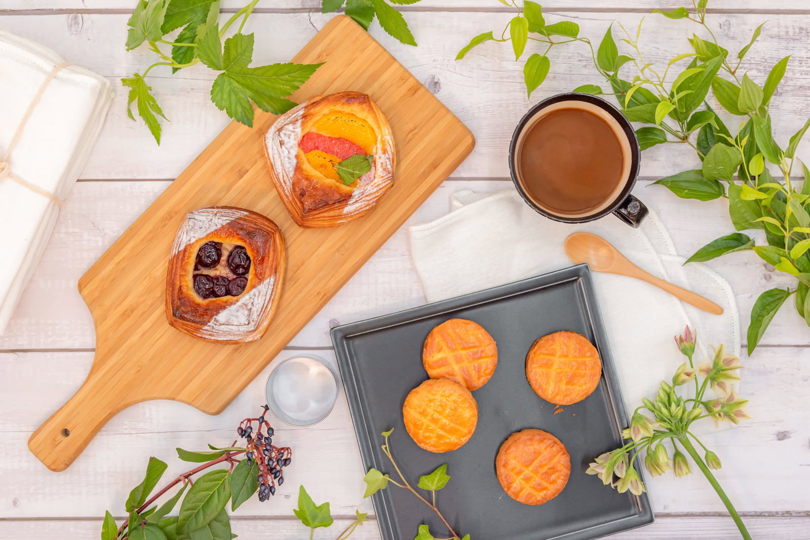 木製まな板の上のフルーツデニッシュとガレットブルトンヌ、コーヒーカップと観葉植物がある朝食シーン