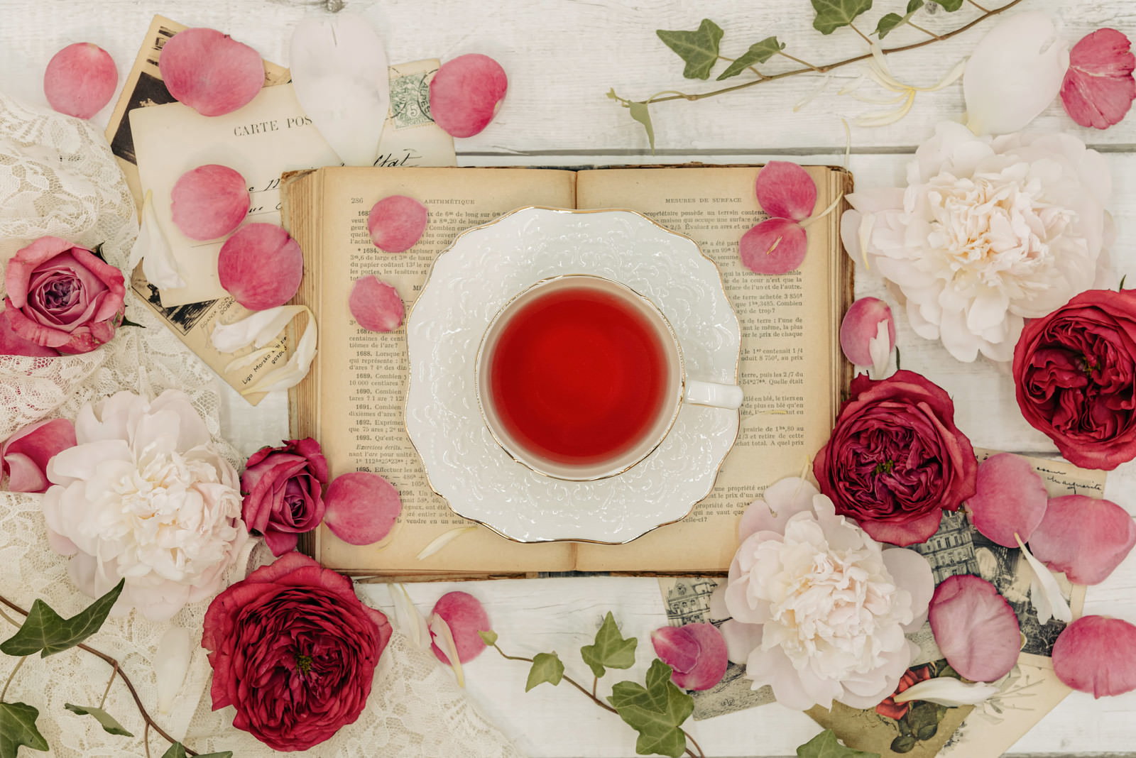 洋書の上に置かれた白いティーカップと赤いローズティー、散らばった薔薇の花びら