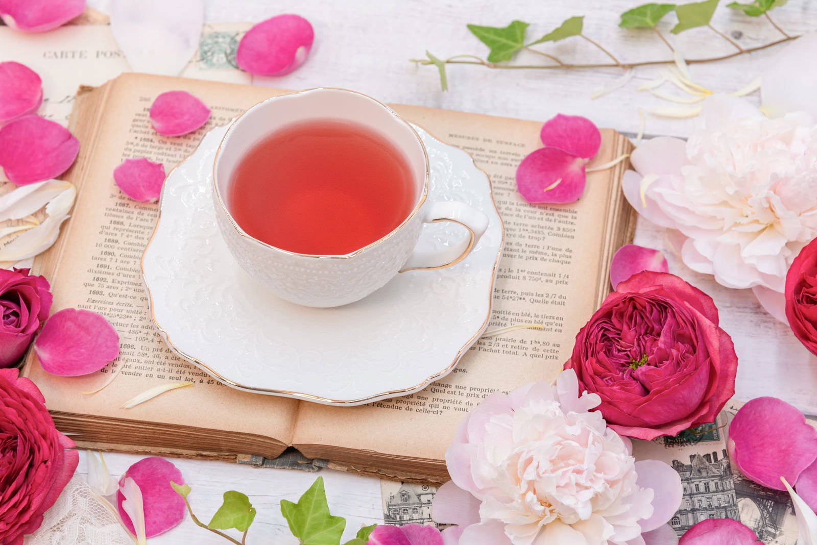 洋書の上に置かれた白いカップのローズティーと散らばったピンクの薔薇の花びら