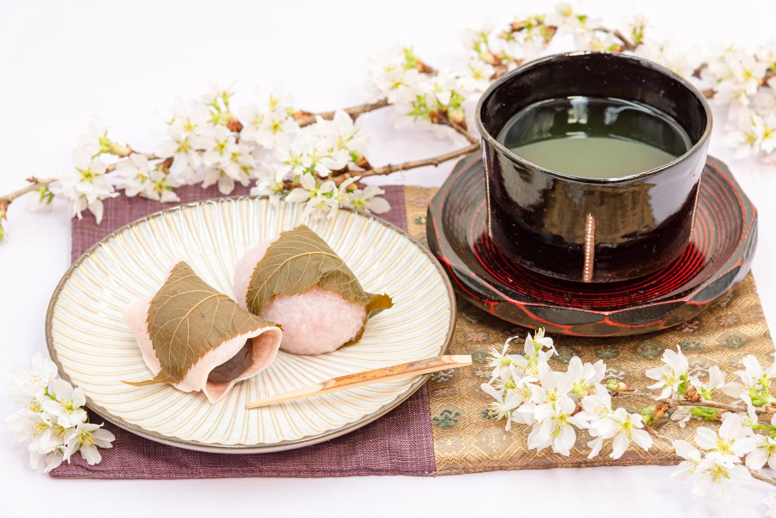 白い器に盛られた桜の葉に包まれたピンク色の桜もちと、黒い碗の抹茶、白い枝花