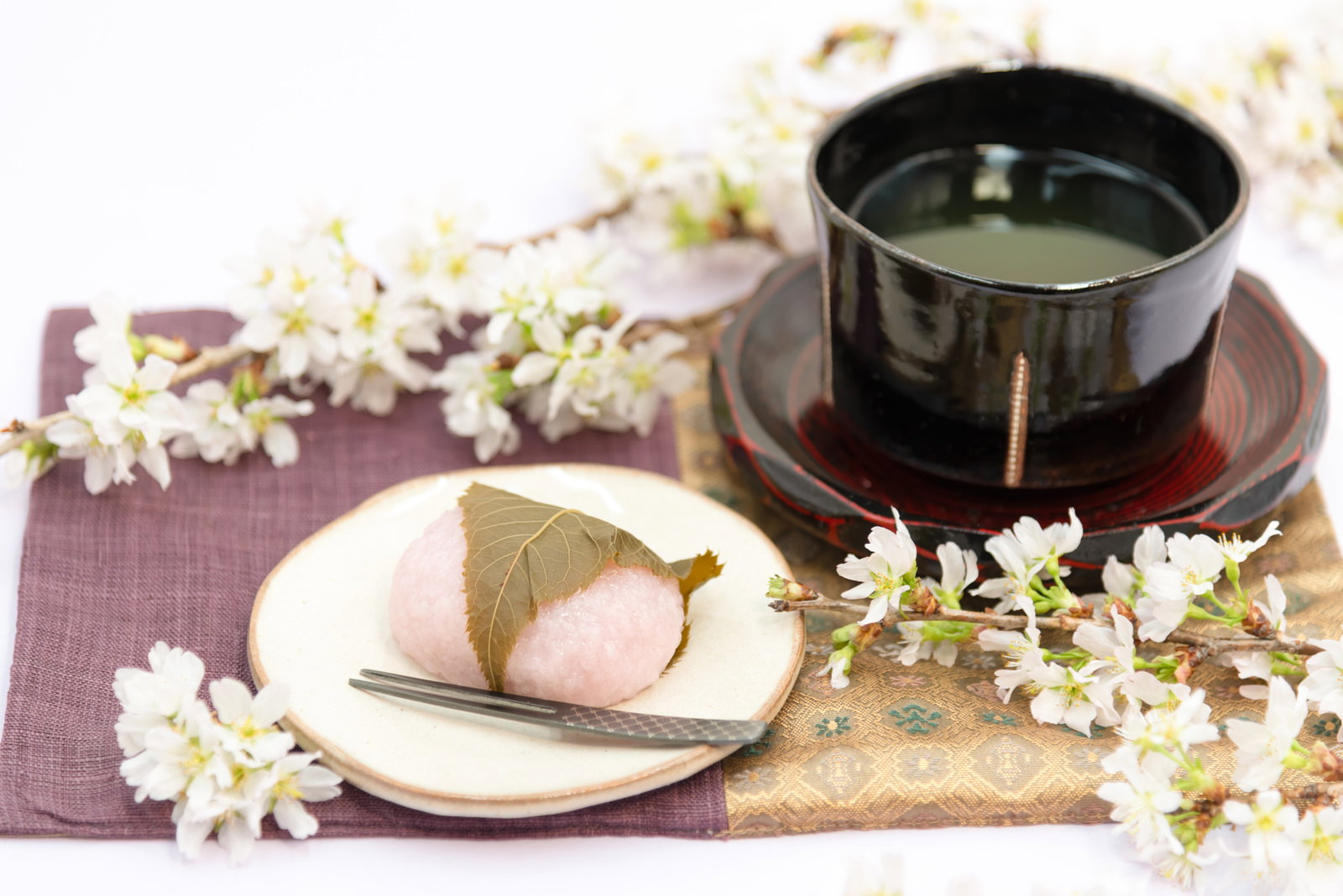 白い皿の桜餅と黒い茶碗の抹茶、桜の花びらが散る和風の茶菓子セット