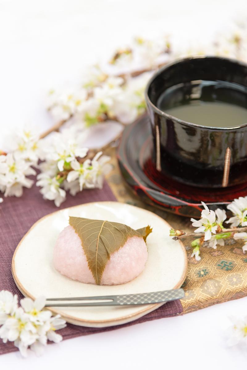 Cherry blossom mochi on a white plate, a bowl of matcha tea, and scattered sakura petals