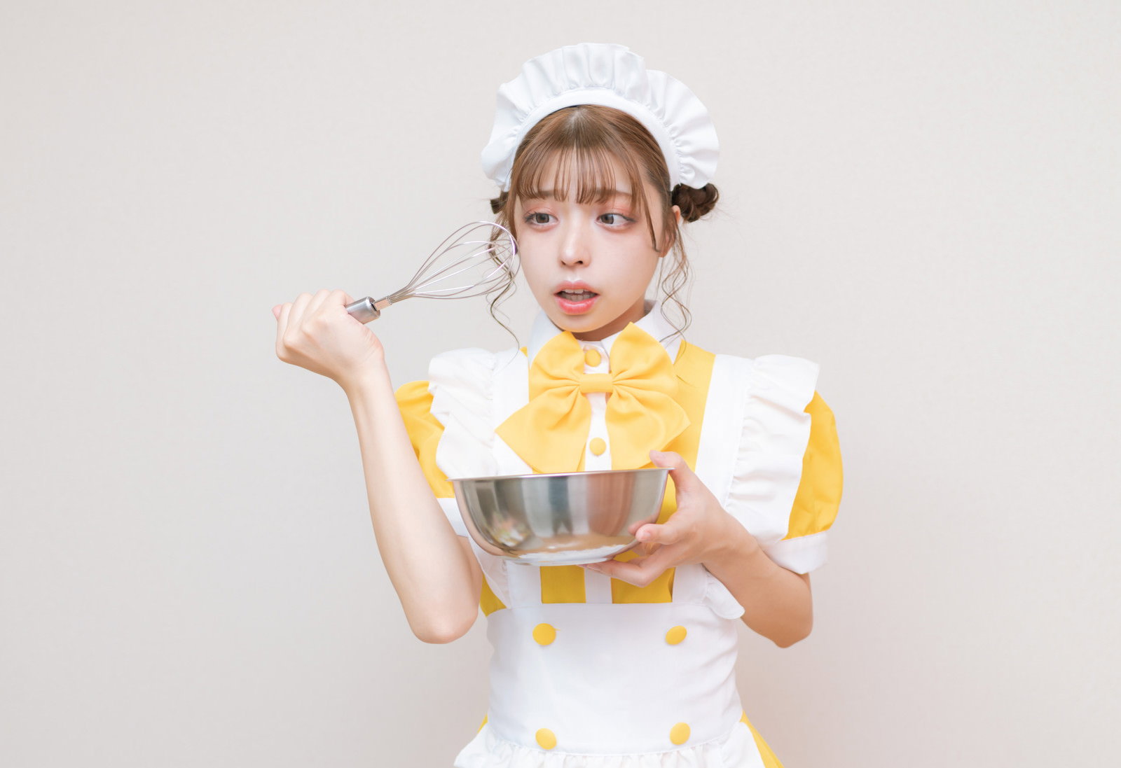 黄色と白のメイド服を着た女性が両手でホイッパーを持ち困惑した表情を見せている［モデル：高尾実生］