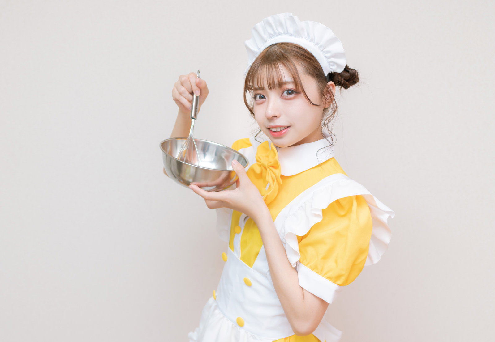 白いエプロンと黄色いリボンのメイド服を着た女性がボールとホイッパーを持つ［モデル：高尾実生］