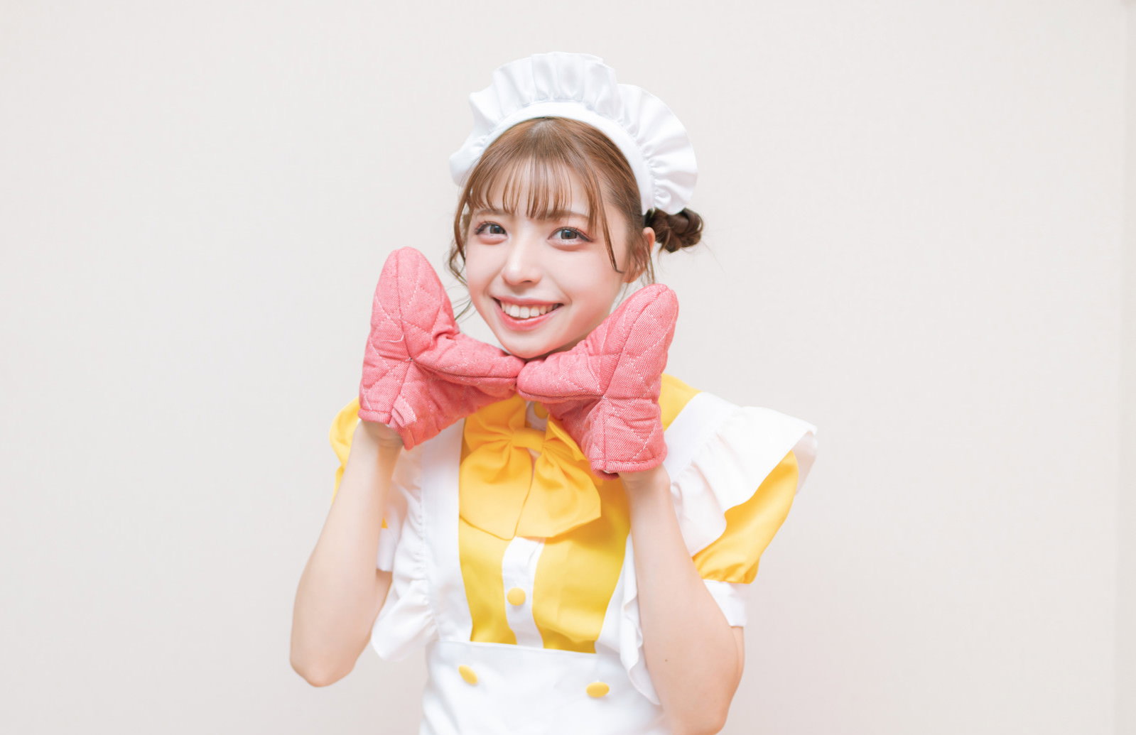 黄色いエプロンドレスを着てミトンを両手に持ち笑顔のメイドコスプレ女性［モデル：高尾実生］