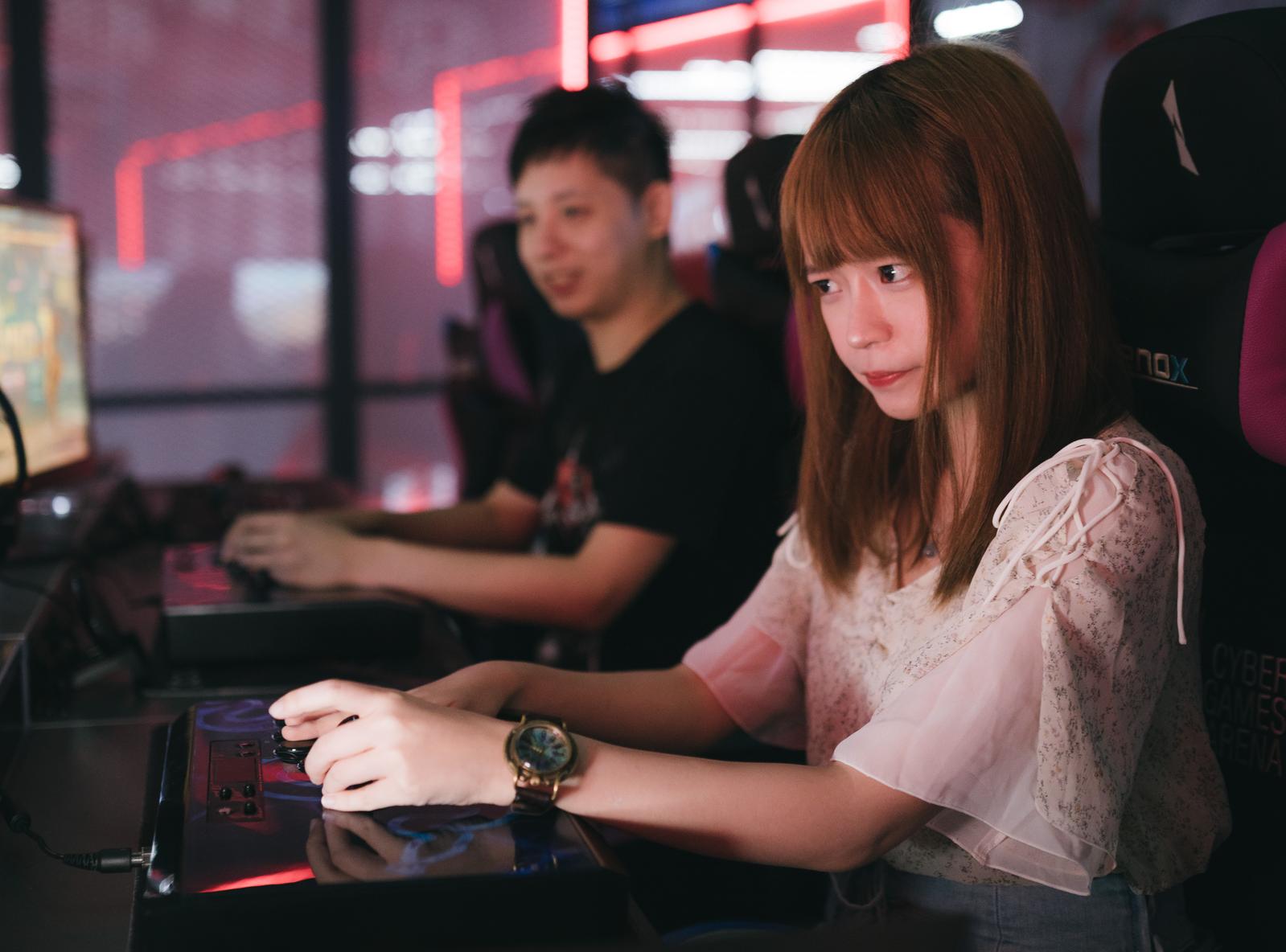 ゲームセンターで格闘ゲームをプレイする女性と隣で見守る男性［モデル：Humanbomb］