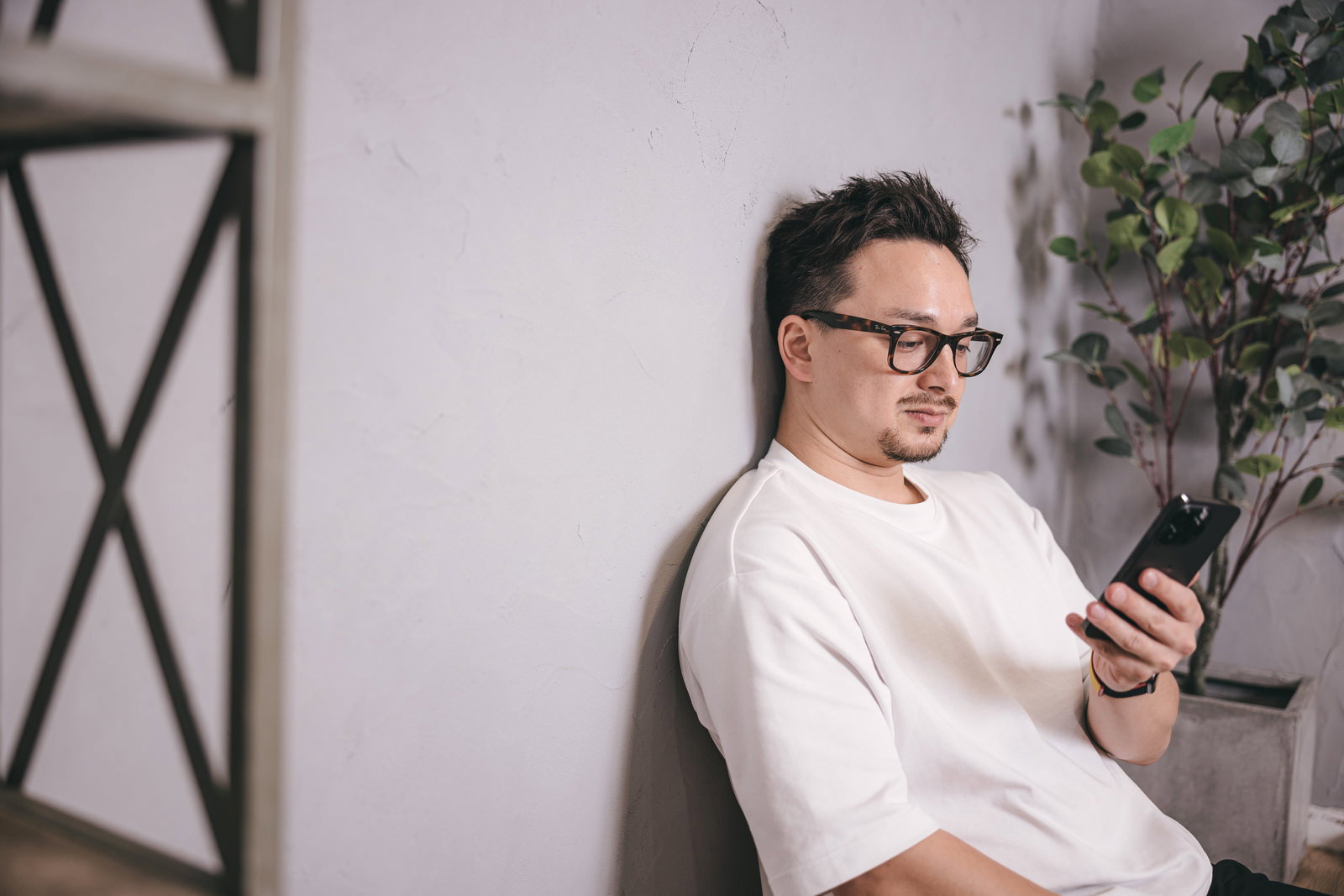 白い壁の前でスマートフォンを見つめる眼鏡をかけた男性［モデル：Max_Ezaki］