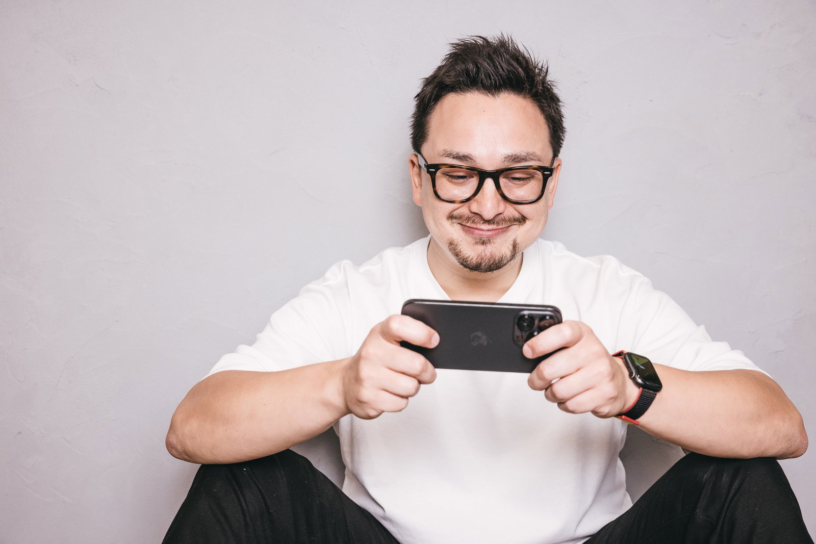 スマートフォンでゲームをしながら笑顔を見せるメガネの男性［モデル：Max_Ezaki］