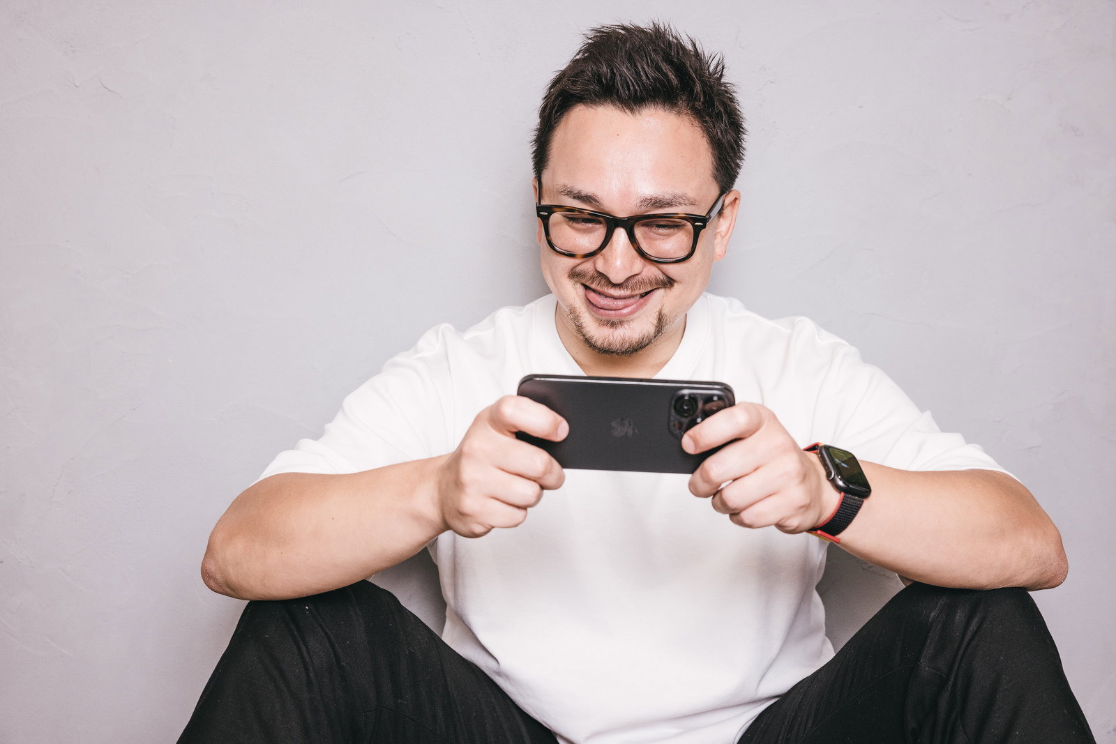 眼鏡をかけた男性が白いTシャツを着てスマホを両手で持ちニヤけている［モデル：Max_Ezaki］