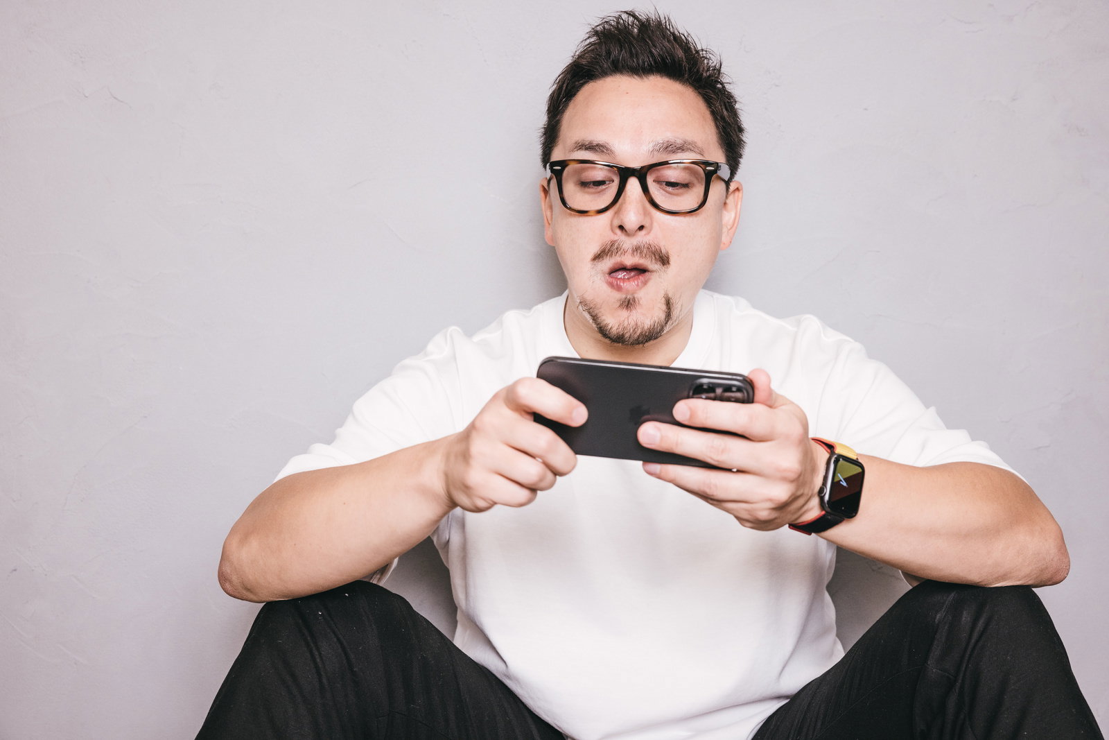 眼鏡をかけた男性がスマートフォンを両手で持ちゲームをプレイしている様子［モデル：Max_Ezaki］