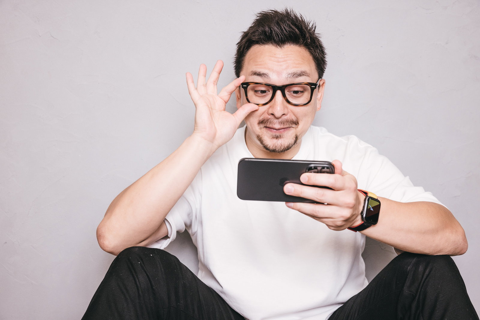 スマホを見ながらOKサインをしてにやける白いTシャツの男性［モデル：ドイツ人ハーフ］