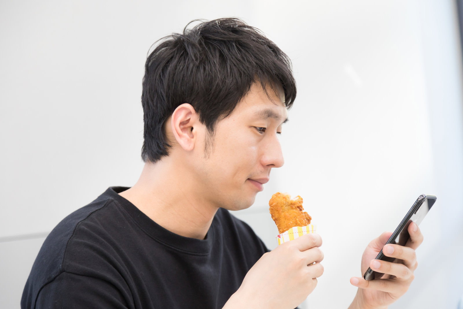 黒いTシャツの男性がフライドチキンを食べながらスマートフォンを見ている［モデル：大川竜弥］