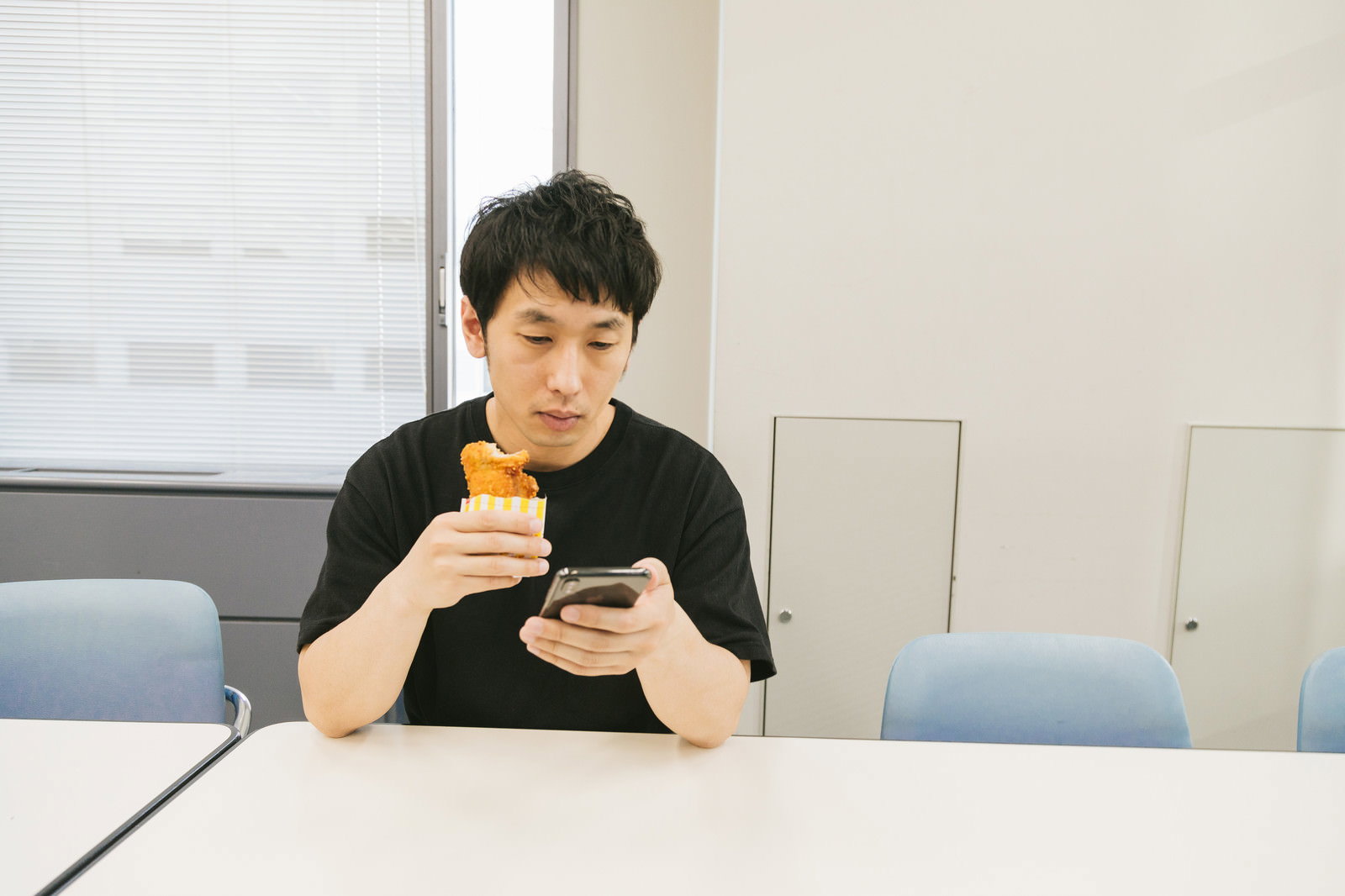会議室でスマートフォンを見ながらフライドチキンを持つ男性［モデル：大川竜弥］