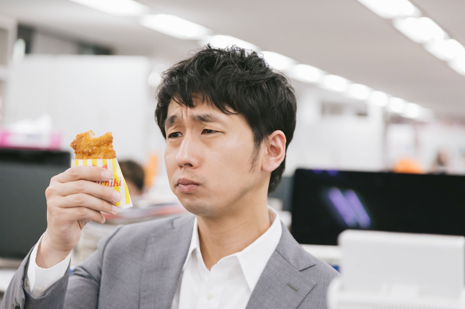 フライドチキンを手に持って困った表情を浮かべるスーツ姿の男性［モデル：大川竜弥］