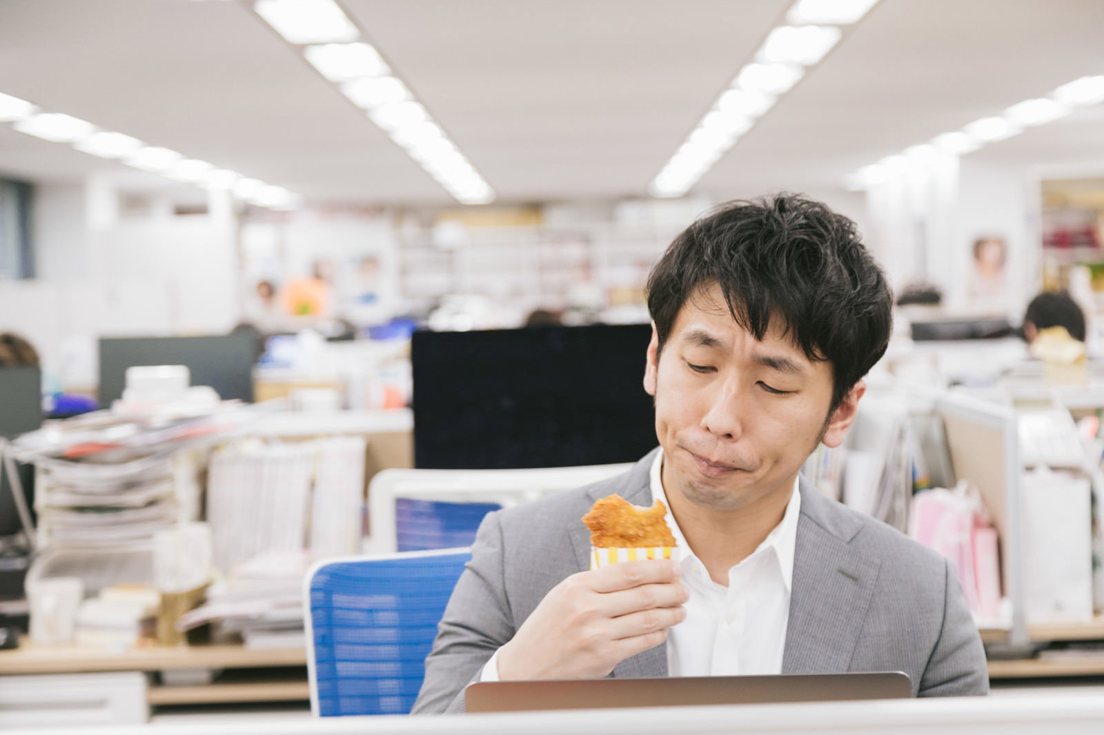 オフィスのデスクで骨なしフライドチキンを食べるスーツ姿の男性［モデル：大川竜弥］
