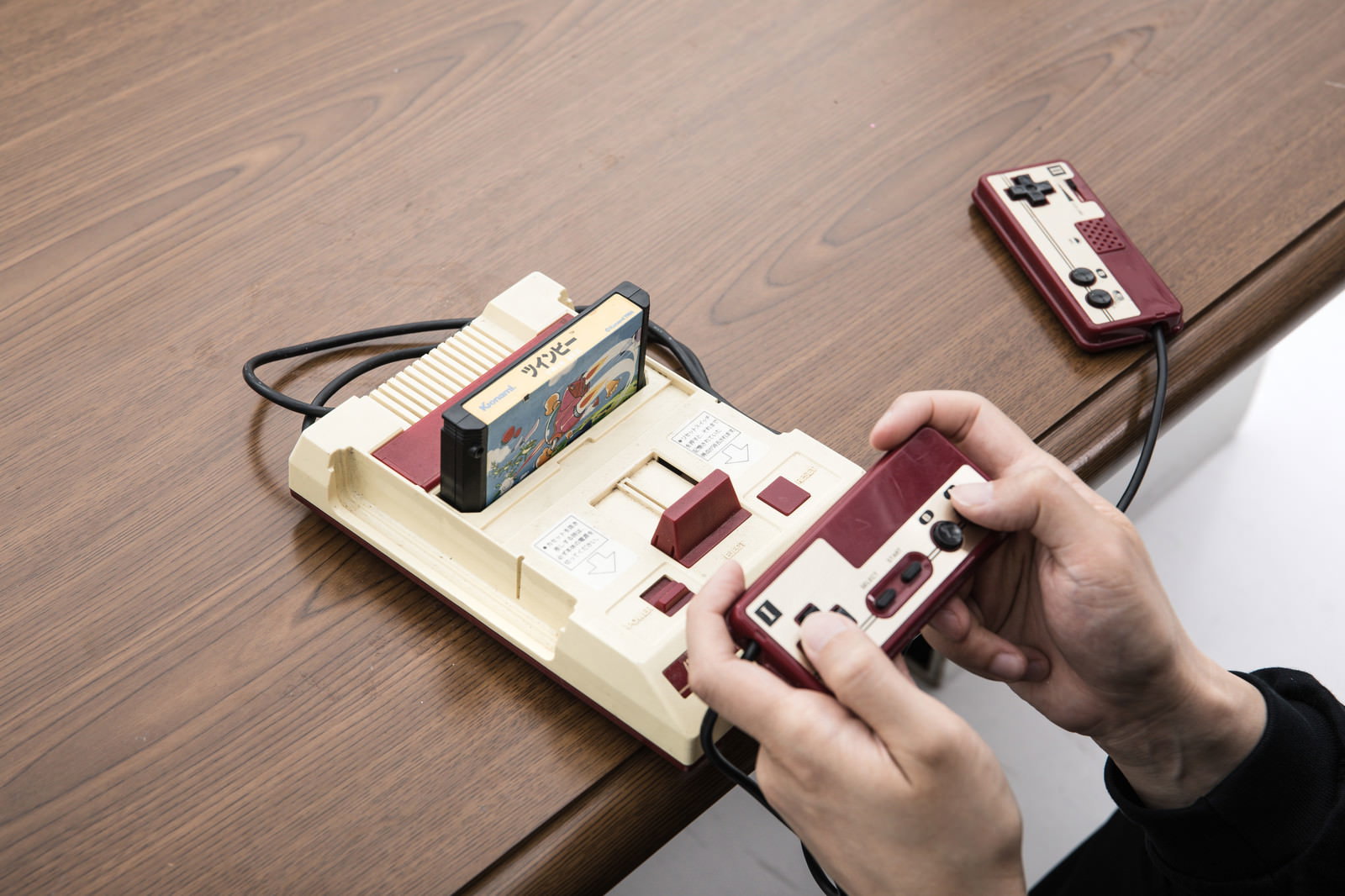 木製テーブルの上でファミコン風ゲーム機のコントローラーを持つ手［モデル：大川竜弥］