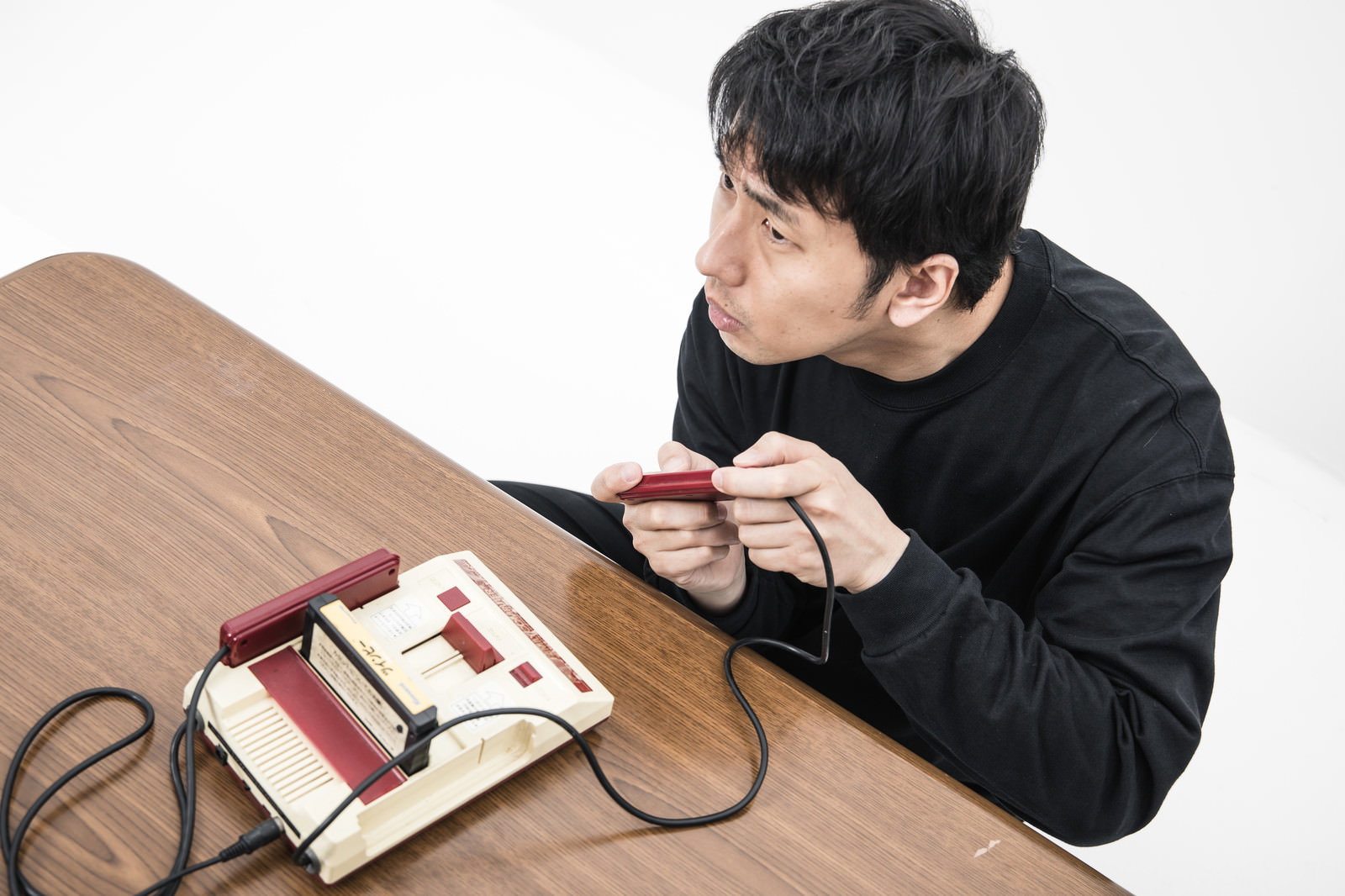 木製テーブルでファミコンのコントローラーを持ってゲームをする黒いスウェット姿の男性［モデル：大川竜弥］