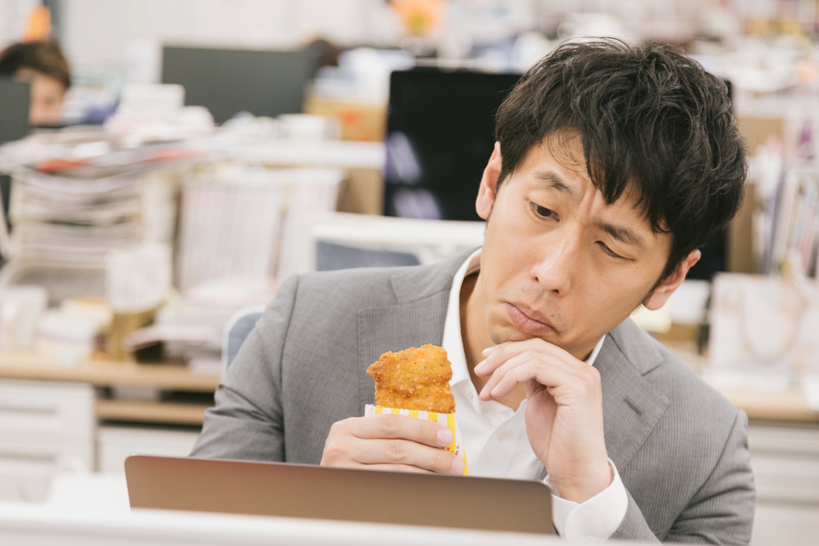 オフィスで食べるかどうか悩みながらフライドチキンを持つスーツ姿の男性［モデル：大川竜弥］