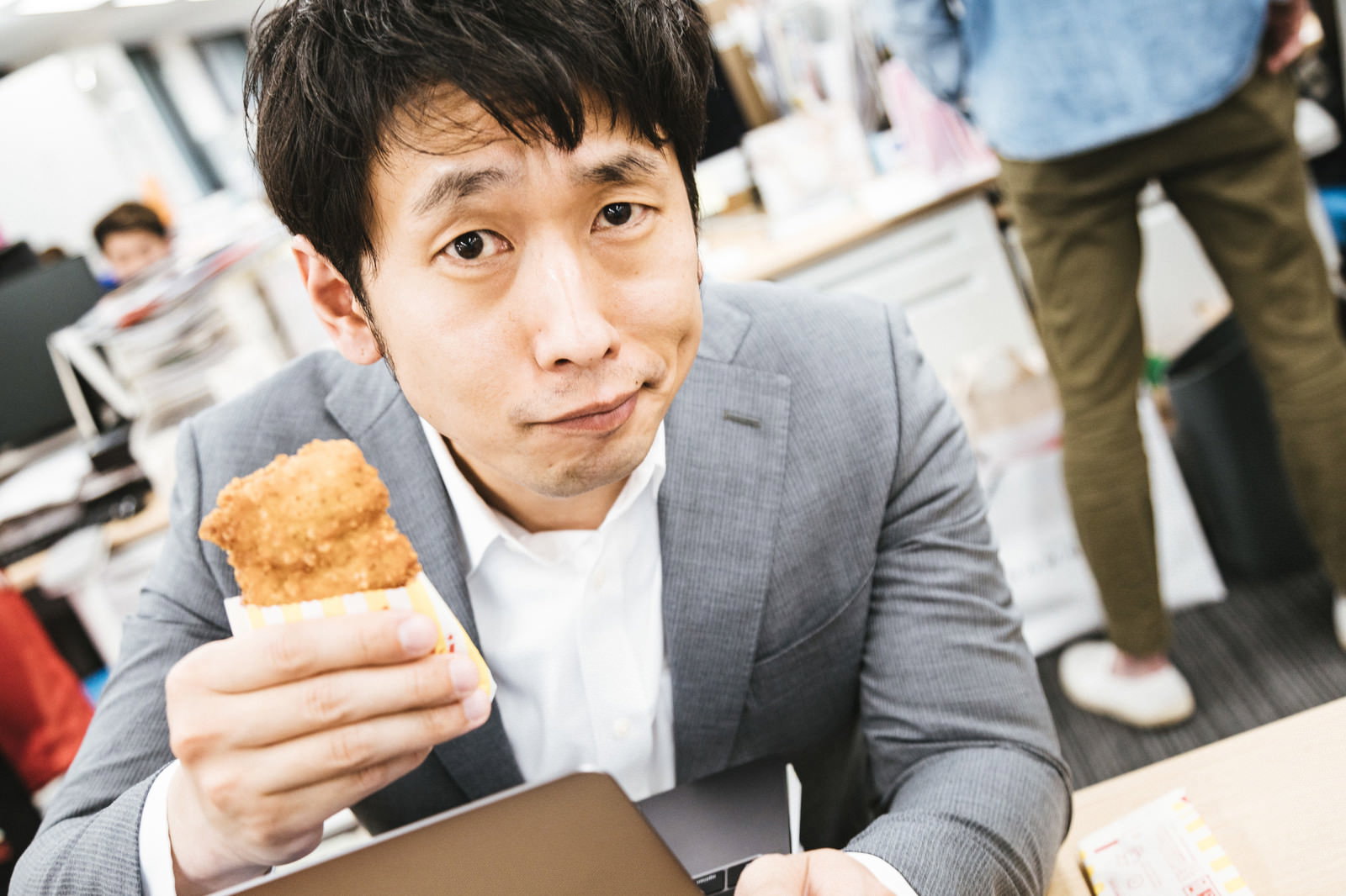 グレーのスーツを着た男性がフライドチキンを手に持って微笑んでいる［モデル：大川竜弥］