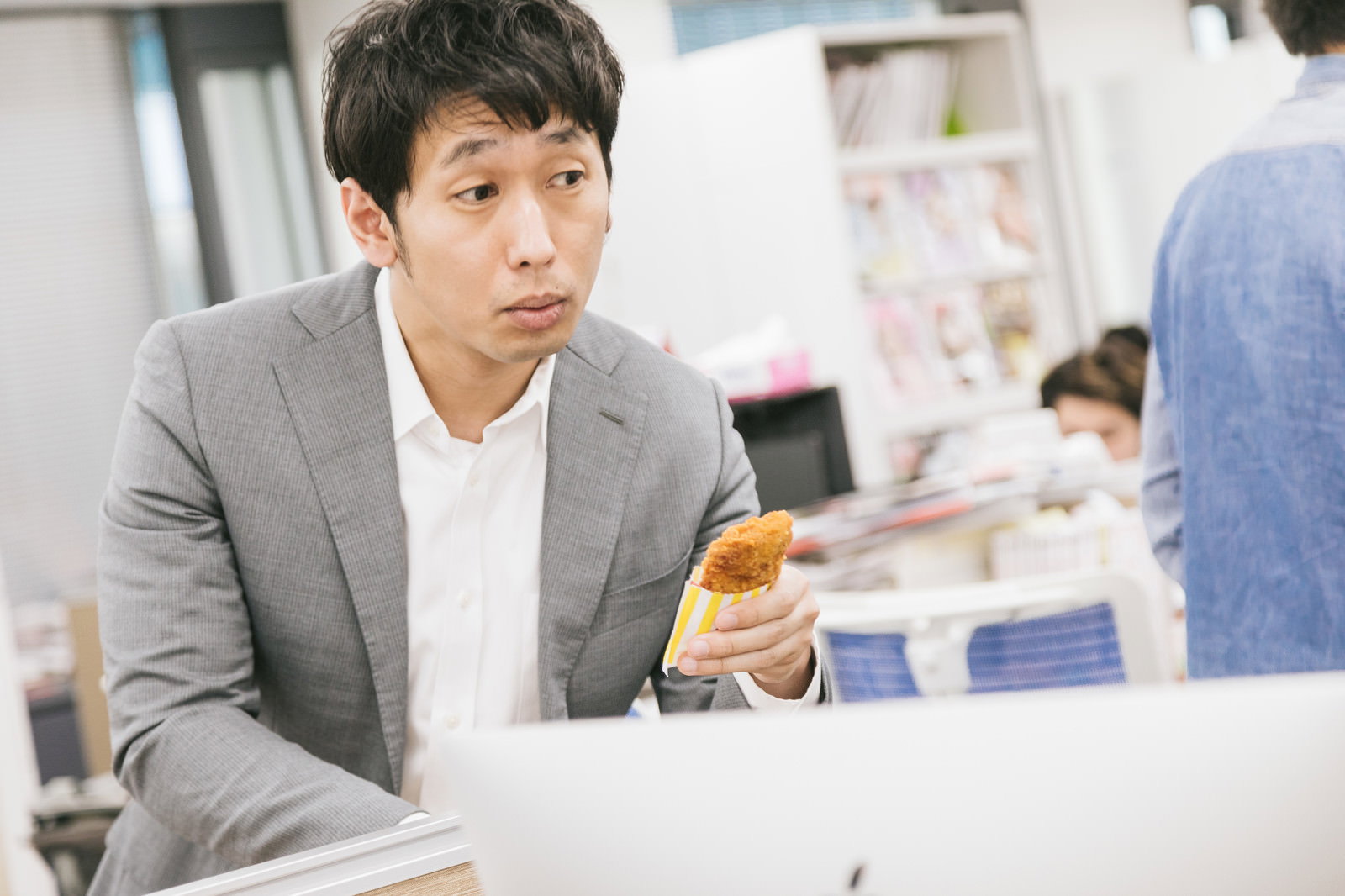 オフィスの机でフライドチキンを手に持ち同僚に勧めるグレースーツの男性［モデル：大川竜弥］