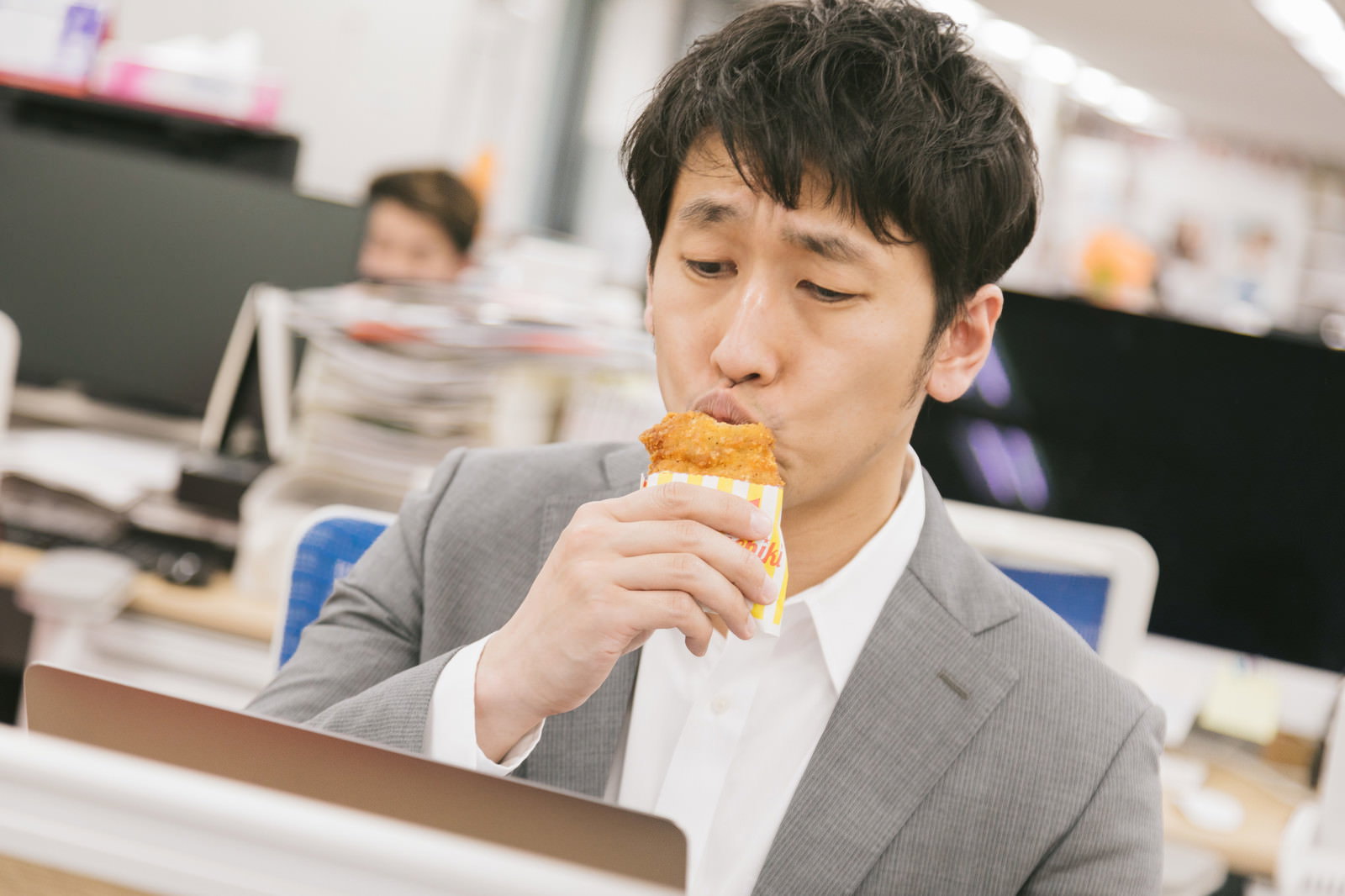 グレーのスーツを着た男性がオフィスでフライドチキンにキスをしている［モデル：大川竜弥］