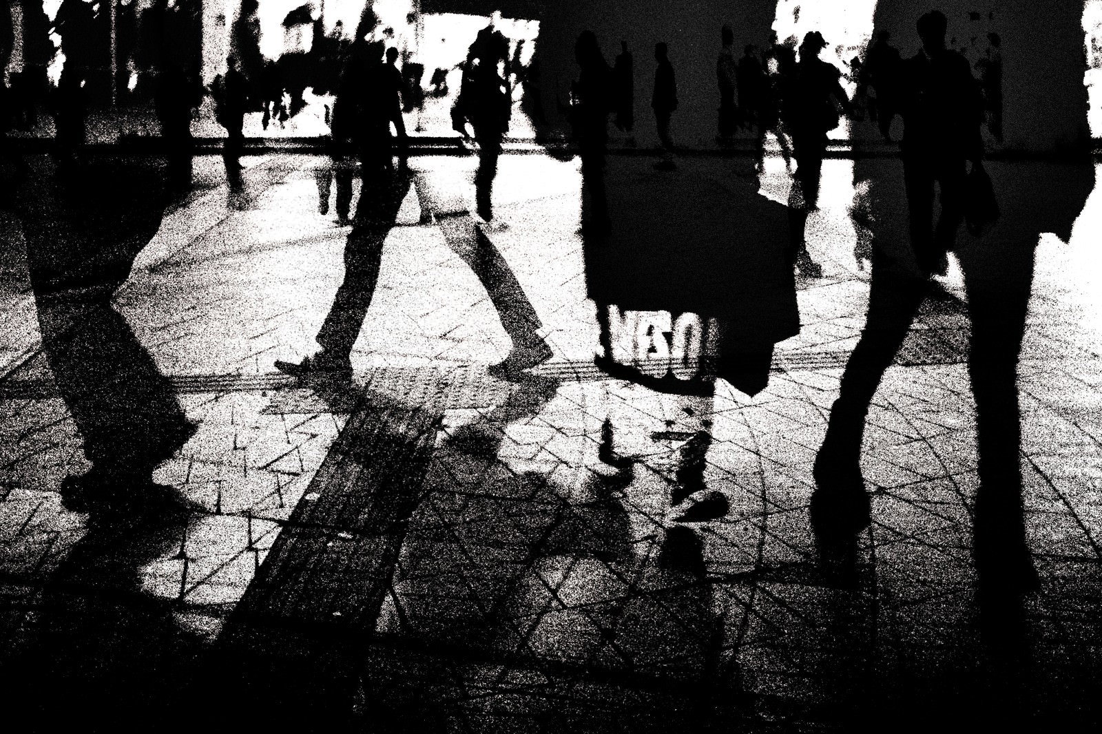 モノクロの高コントラスト写真。白い路面に複数の人影が映り込み、通行人のシルエットが交差する人混みの風景