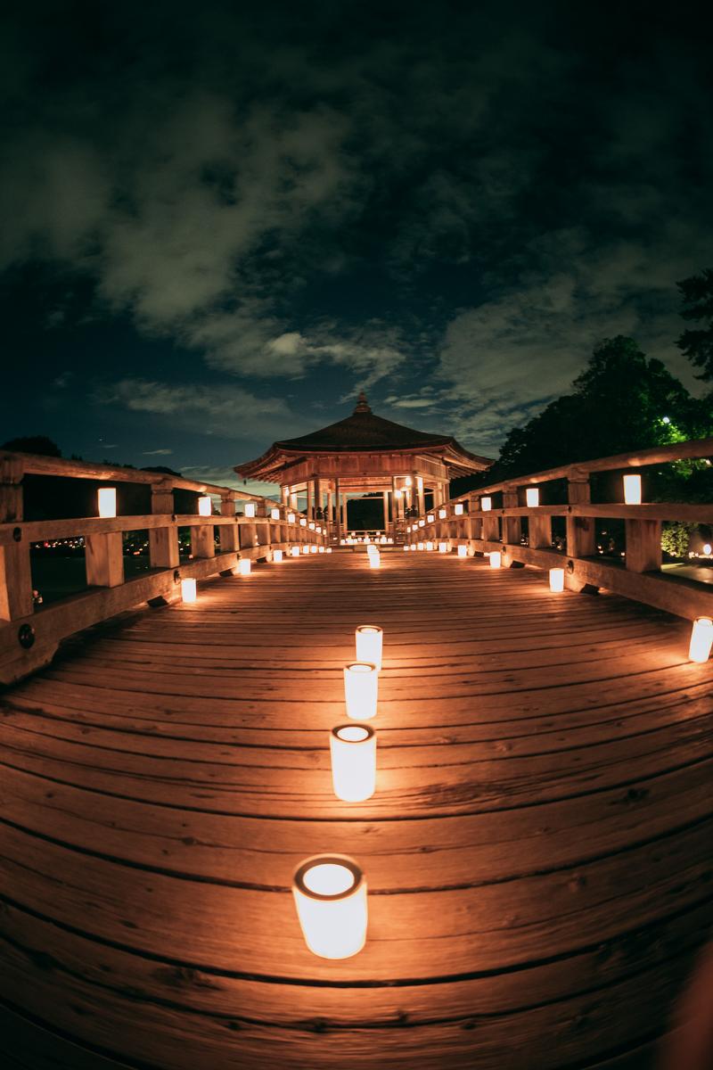 奈良の燈花会で、八角形の建築物を背景に橋に並ぶ多くのろうそくが夜間にライトアップされている光景