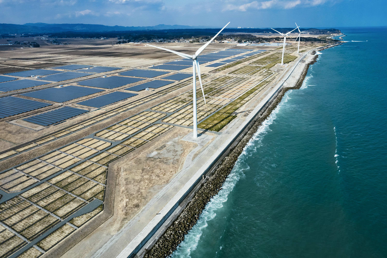 南相馬市鹿島区の海岸に設置された太陽光パネルと風力発電機の空撮写真。海側には白い風車が3基立ち、手前には青いソーラーパネルが格子状に配置されている
